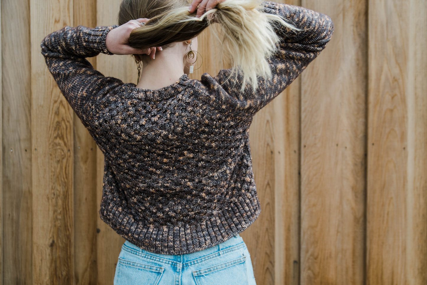 See from behind, Haley pulls her long blonde hair up of her shoulders to show off the ribbed collar and raglan construction of her sweater. The sweater is cropped, falling at her waist, and features a honeycomb stitch pattern on the torso.