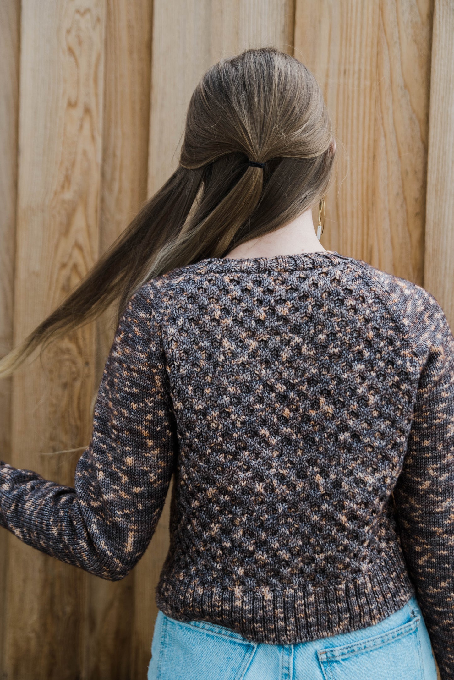 Haley adjusts her blonde hair, seen from behind. She wears a variegated brown sweater, knit with a honeycomb pattern on the torso, with blue jeans.