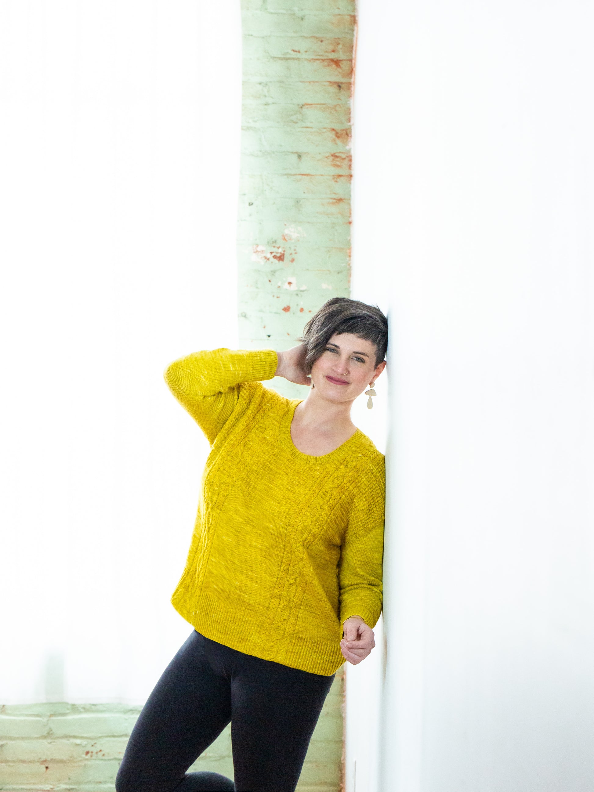 Jen leans against a white wall, smiling at the camera. She wears a knit pullover, made from yellow yarn and featuring a cable and seed stitch pattern.