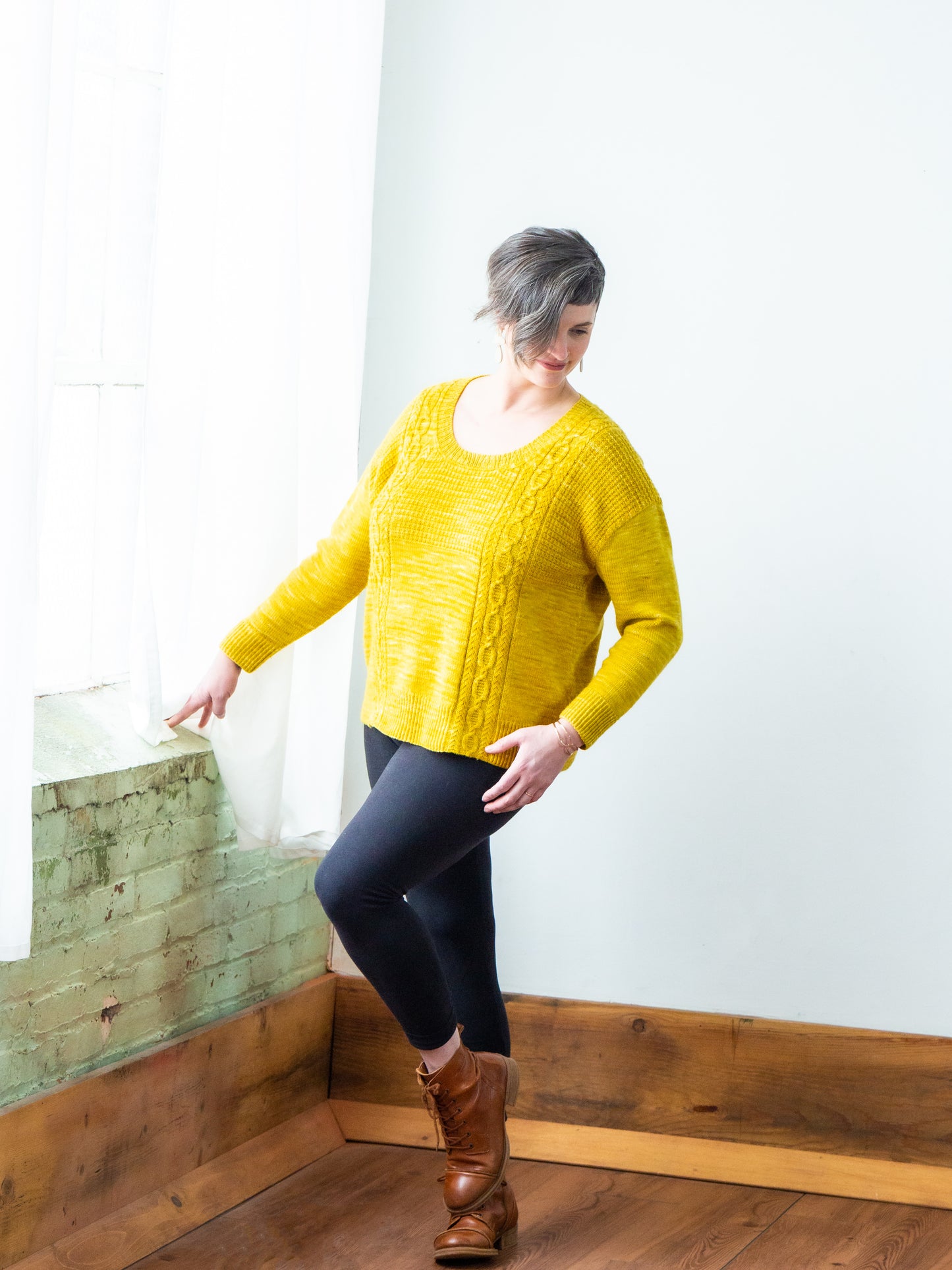 Jen wears a bright yellow sweater with blue pants and brown boots. The sweater is knit with a scoop neck and cabled strips.