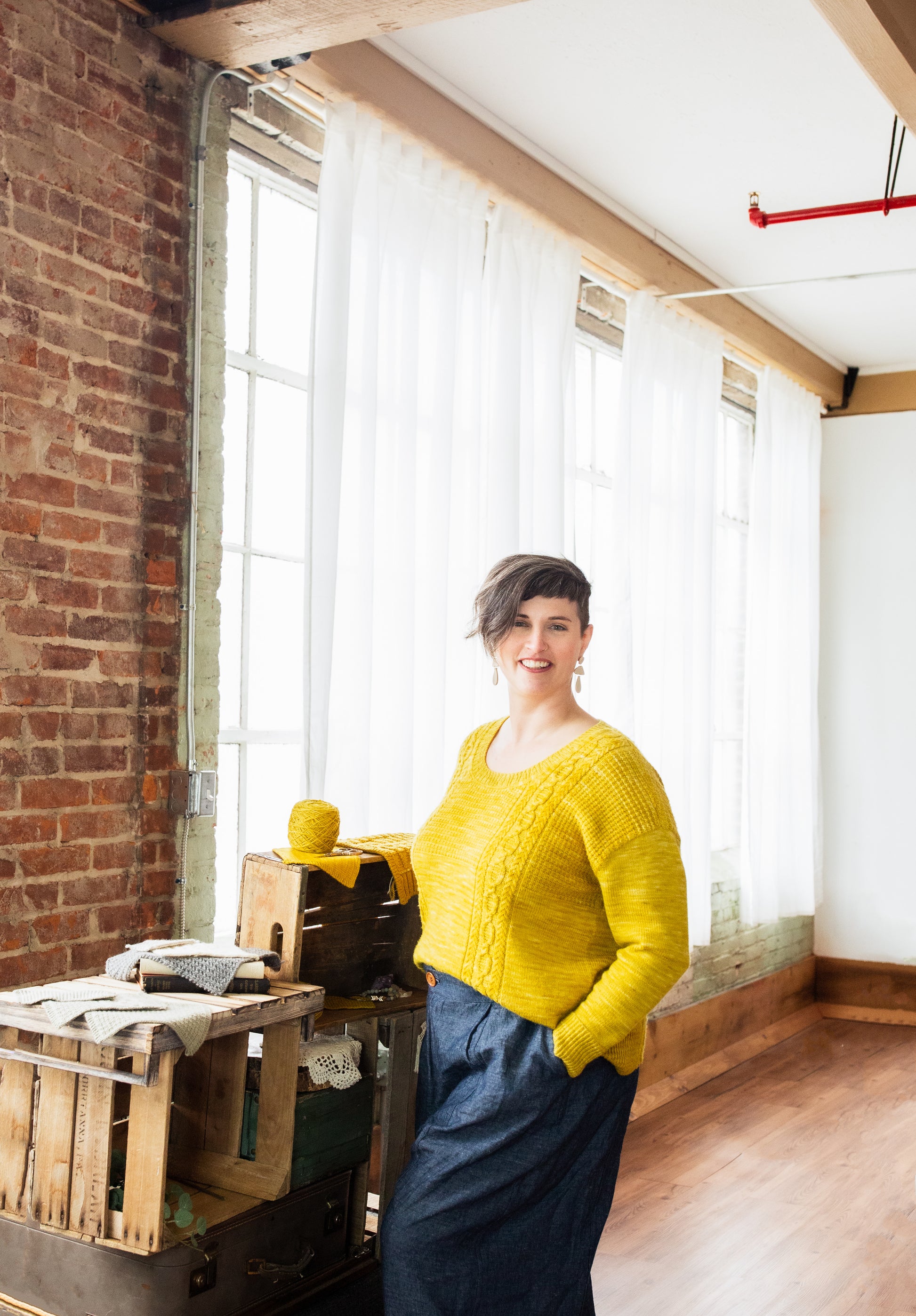 Seen at a 3/4 angle, Jen wears a bright yellow sweater with blue pants. The sweater is knit with a scoop neck and a cable and seed stitch design.
