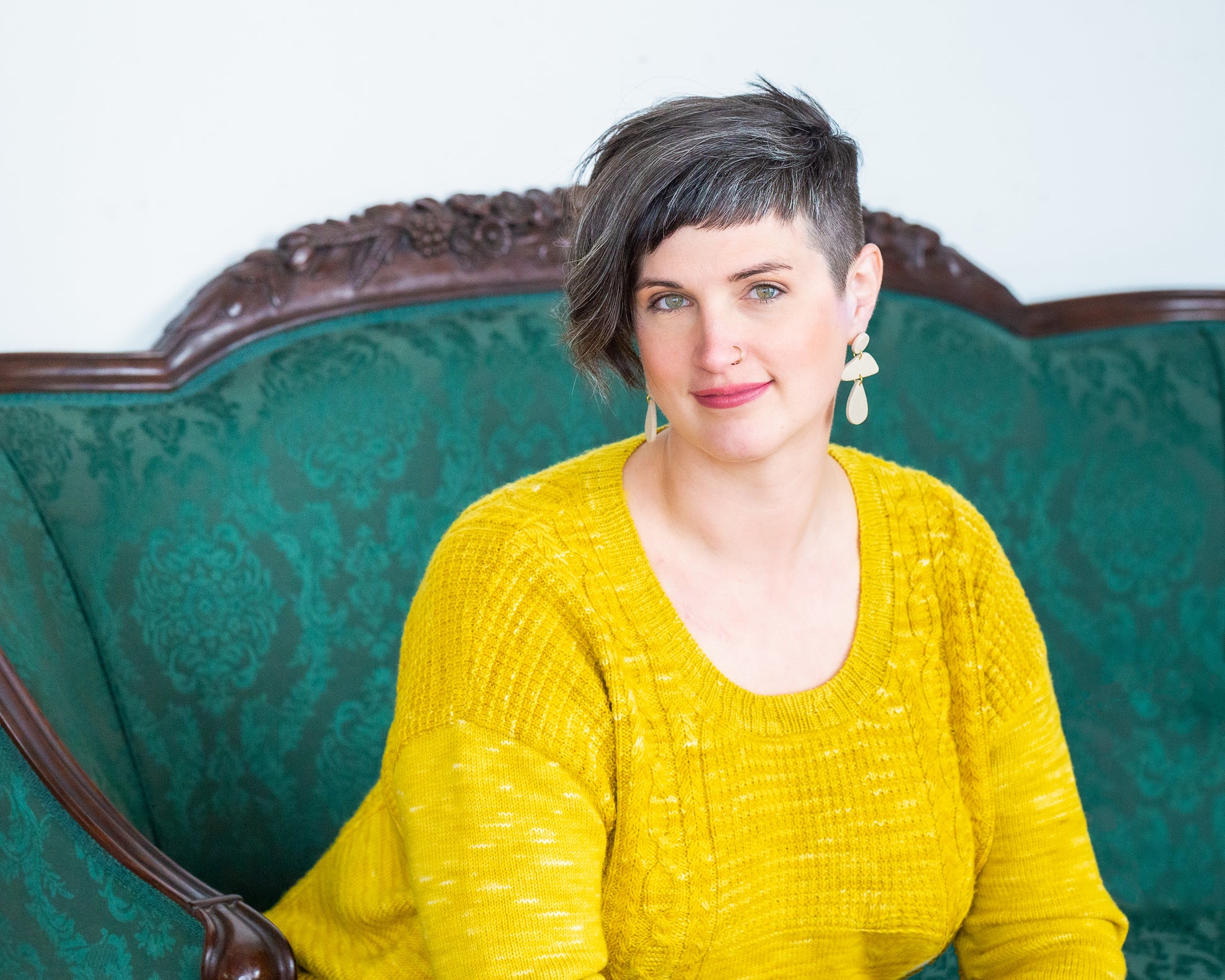 Jen sits on a green couch, leaning forward and smiling at the camera. She wears a bright yellow pullover, featuring a scoop neck and knit with a cable and seed stitch design.