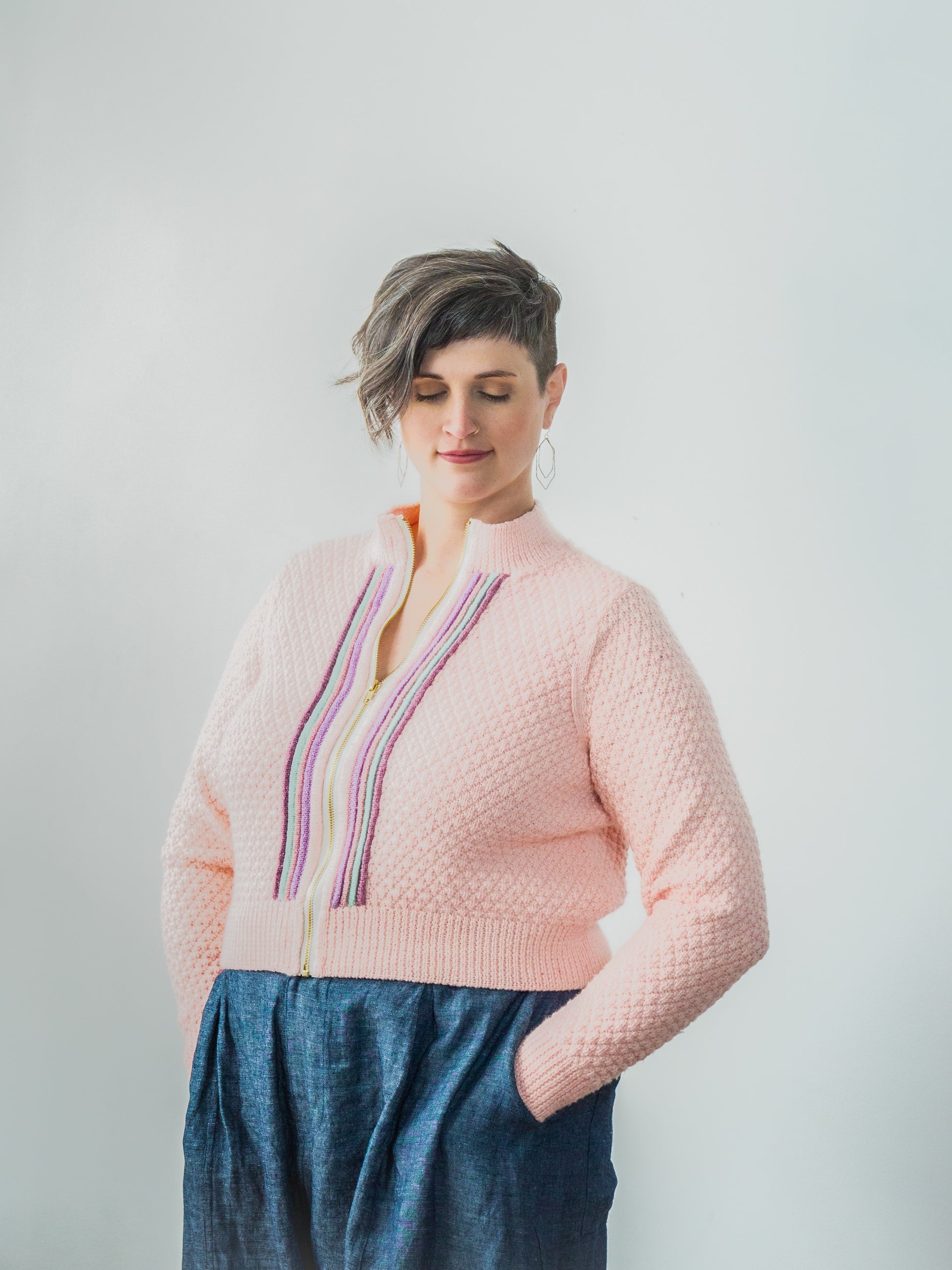 Jen, hands in the pockets of a blue skirt, wears a light pink moss stitch jacket. The jacket features a gold zipper, with multi-colored plackets around the zipper band.