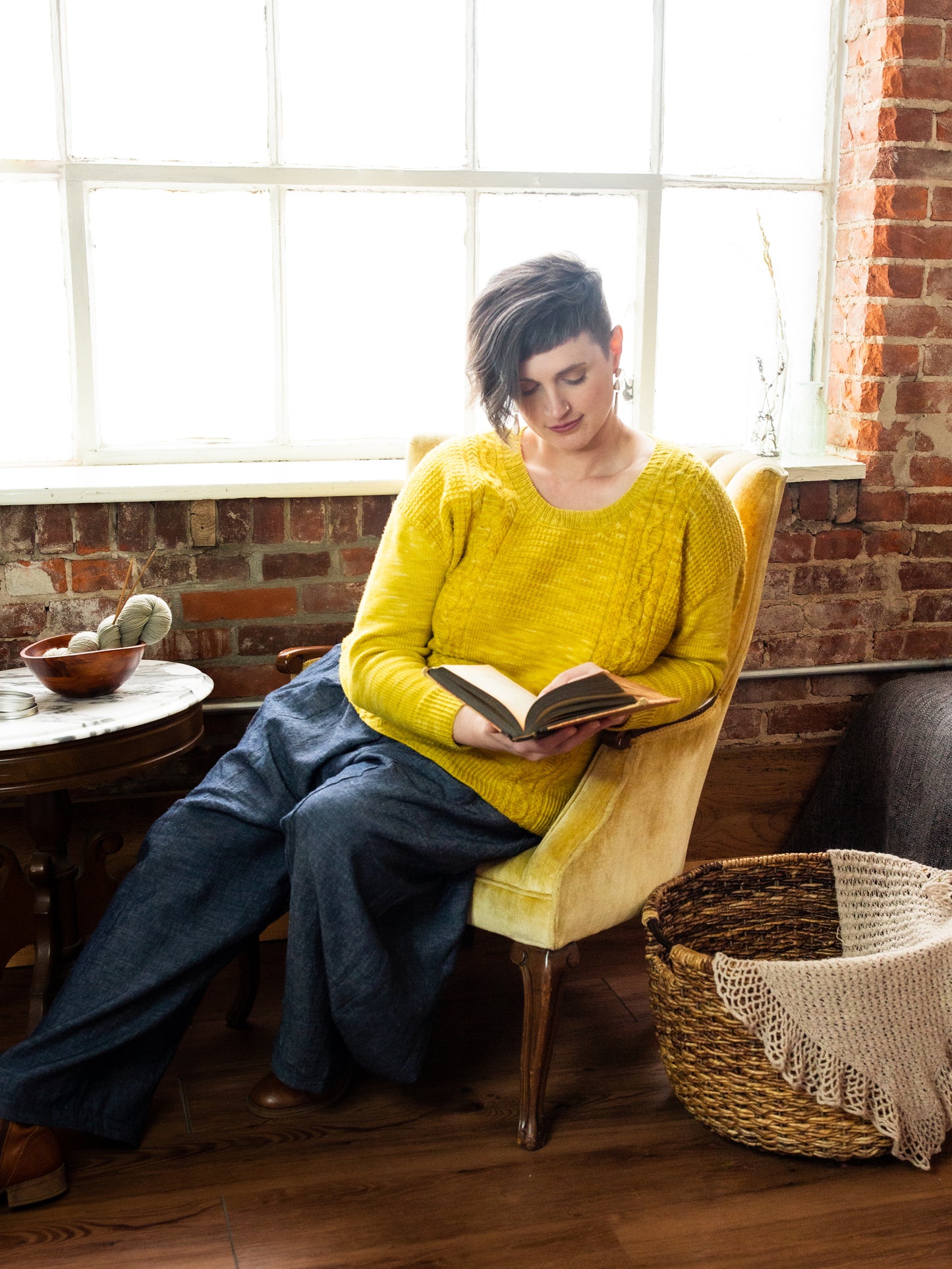 Jen reclines in a chair in front of a window, reading a book. She wears a bright yellow hand knit pullover, with wide leg blue pants and brown boots.