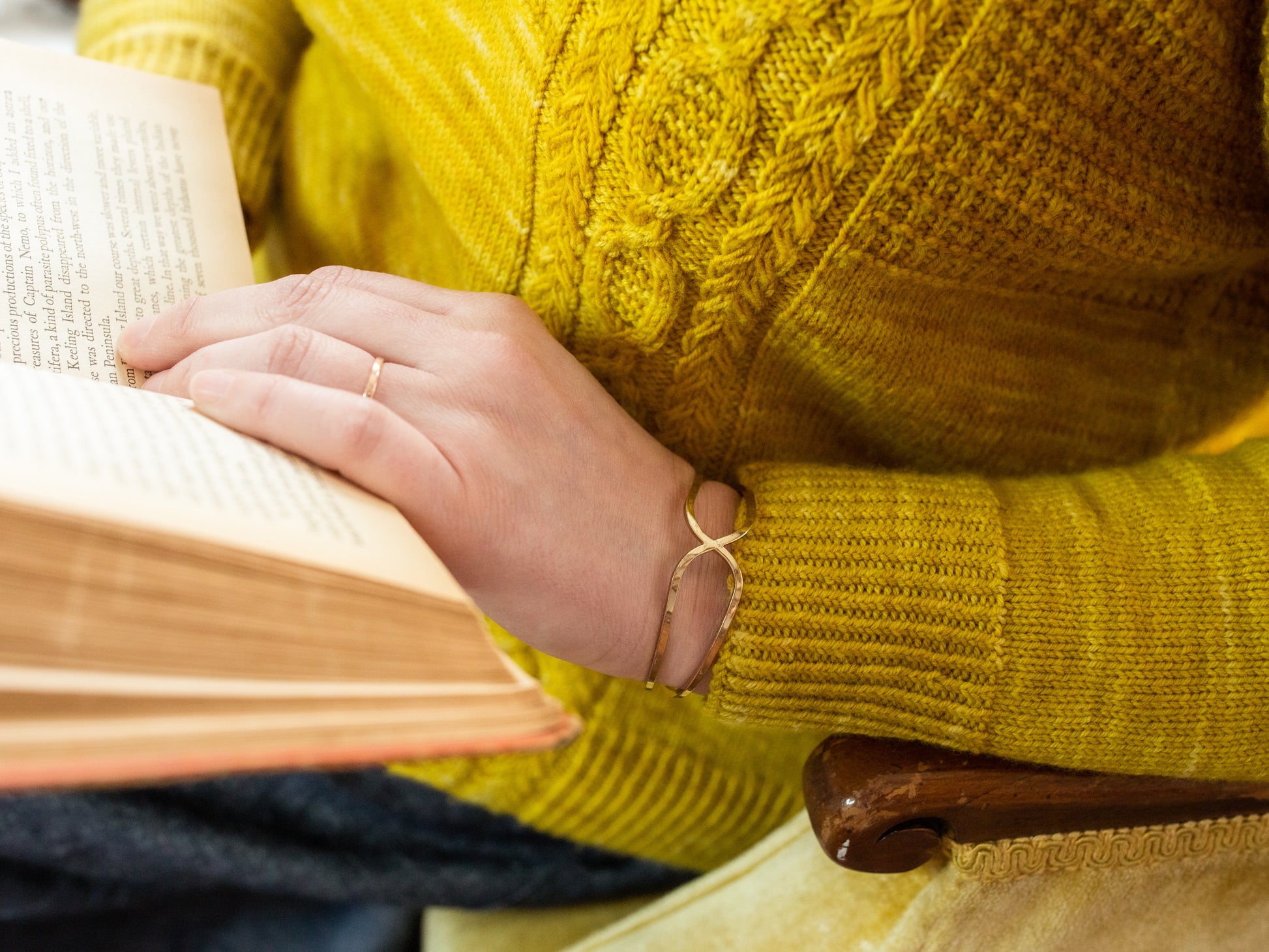 Seen close up, Jen reads a book while wearing a bright yellow sweater. The camera focuses on the ribbed cuff and cable sections of the sweater.