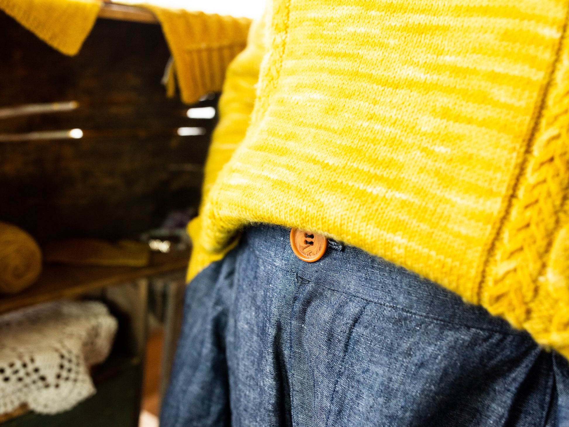 Seen close up, Jen wears a bright yellow sweater, knit with cable strips, tucked into a pair of blue pants. Knit swatches can be seen in the background on a wooden shelf.