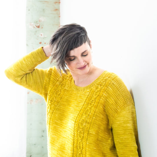 Jen leans against a white wall, one hand in her hair. She wears a bright yellow pullover, knit with two cables stripes down the front.