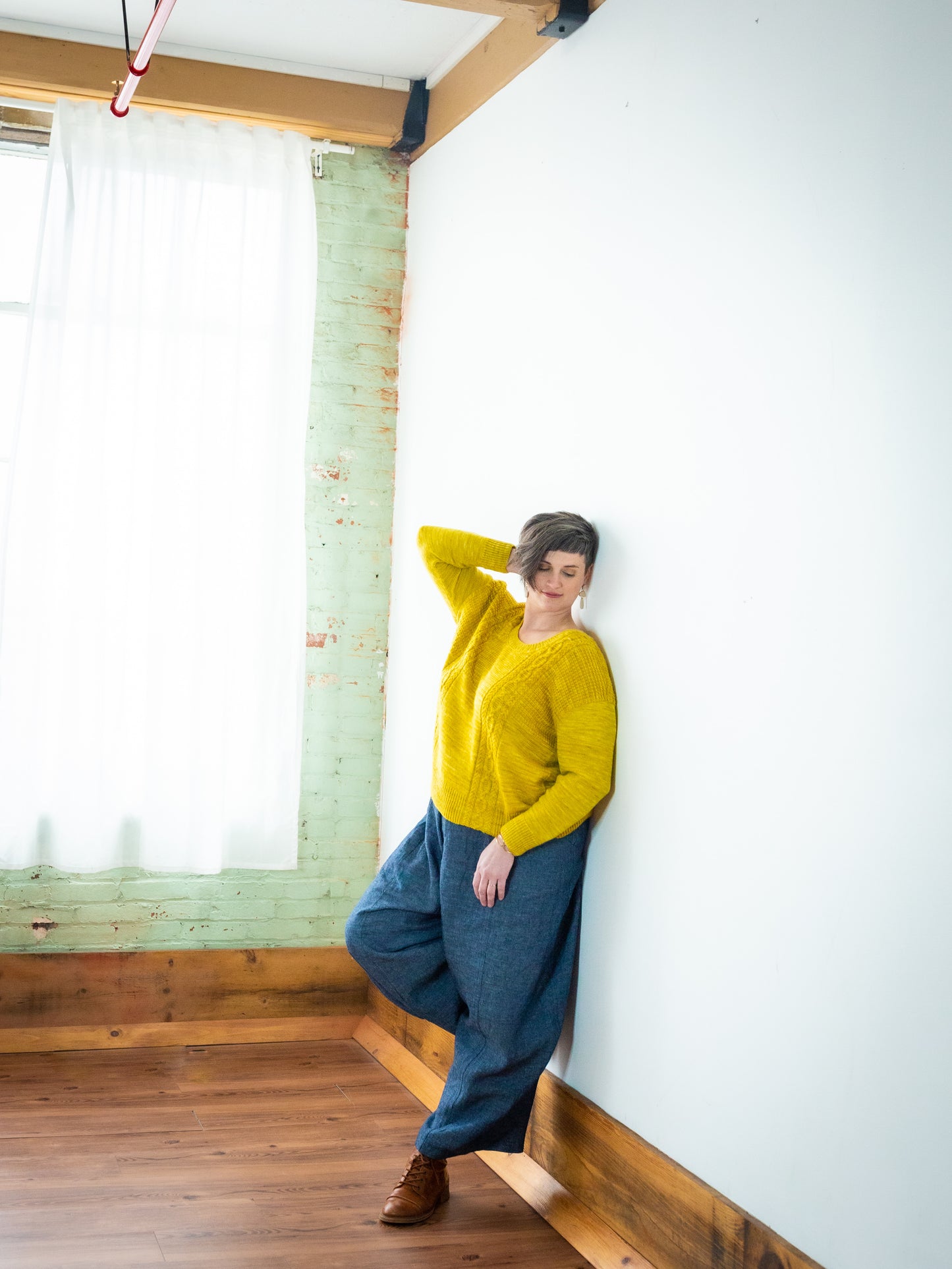 Jen leans against a white wall, wearing brown boots and blue wide leg pants with a yellow pullover. The sweater is knit with cable details and a scoop neck.