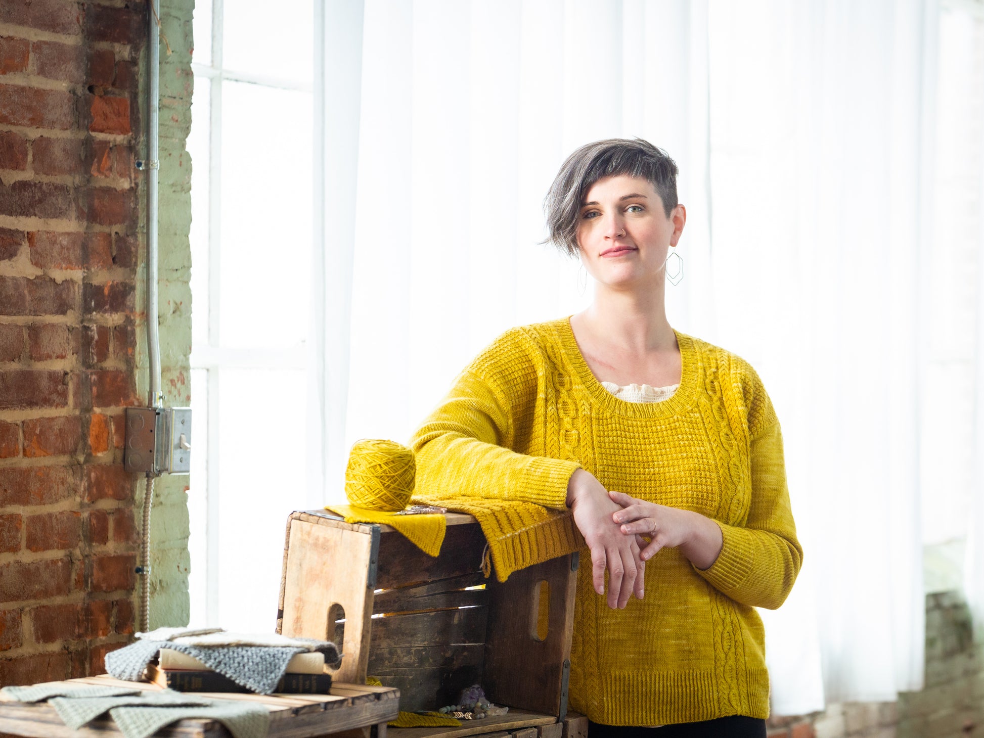 Jen leans against a wooden box, smiling at the camera. She wears a hand knit, bright yellow sweater. The scoop neck shows a white camisole underneath. A matching ball and swatch of yellow yarn can be seen.