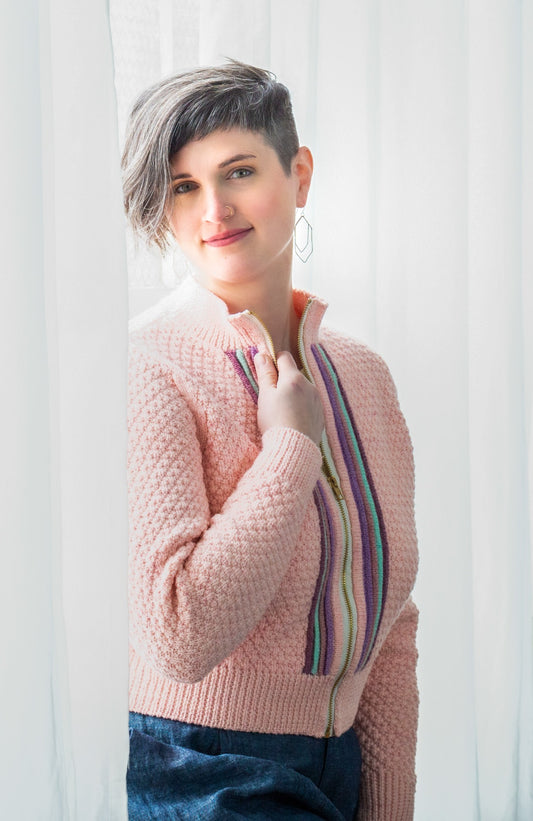 Jen, seen from a 3/4 angle, smiles at the camera. She wears a light pink knit jacket, designed with a moss stitch pattern and featuring knit plackets around the zipper.