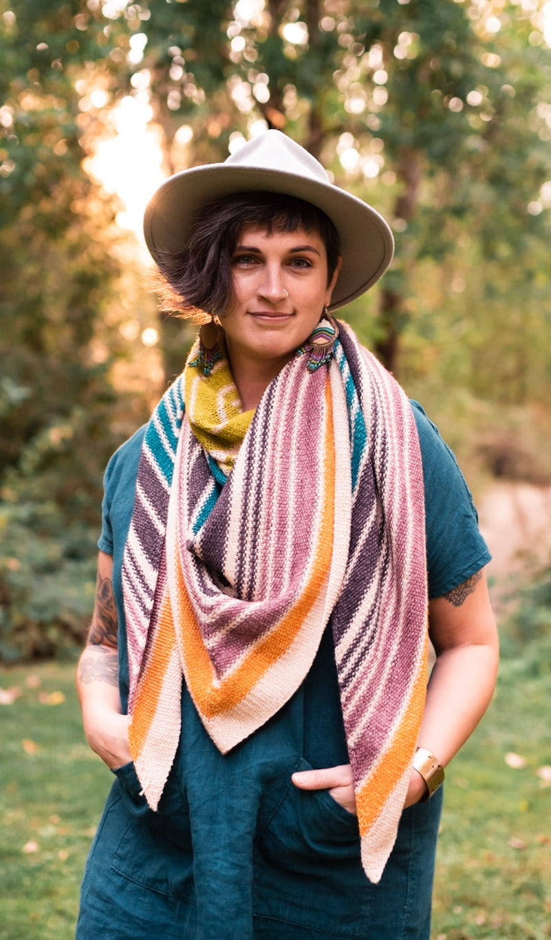 Jen, standing outside, smiles at the camera. She wears a blue dress and hits hat with a striped, hand knit shawl.