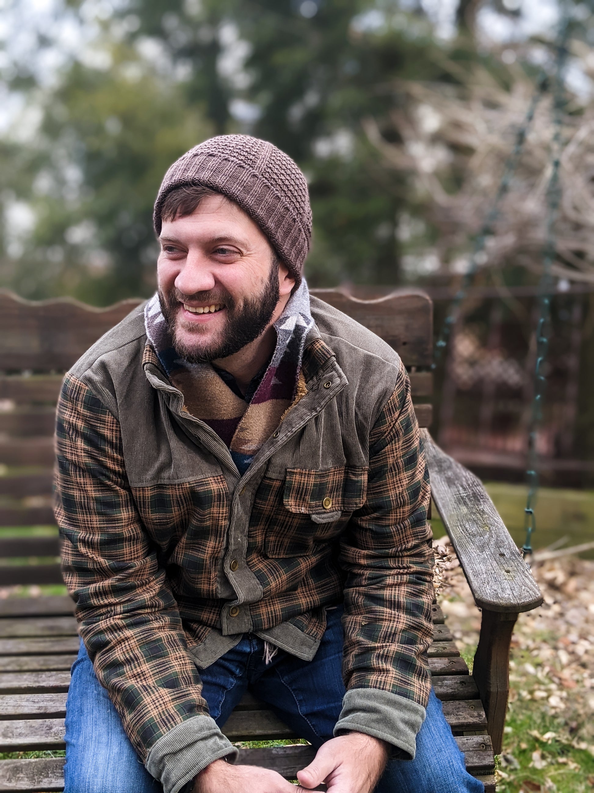Drew, a man with a dark beard, sits outside on a bench. He wears a brown beanie, knit with a ribbed edge and a seed stitch design, with a brown coat and blue jeans.