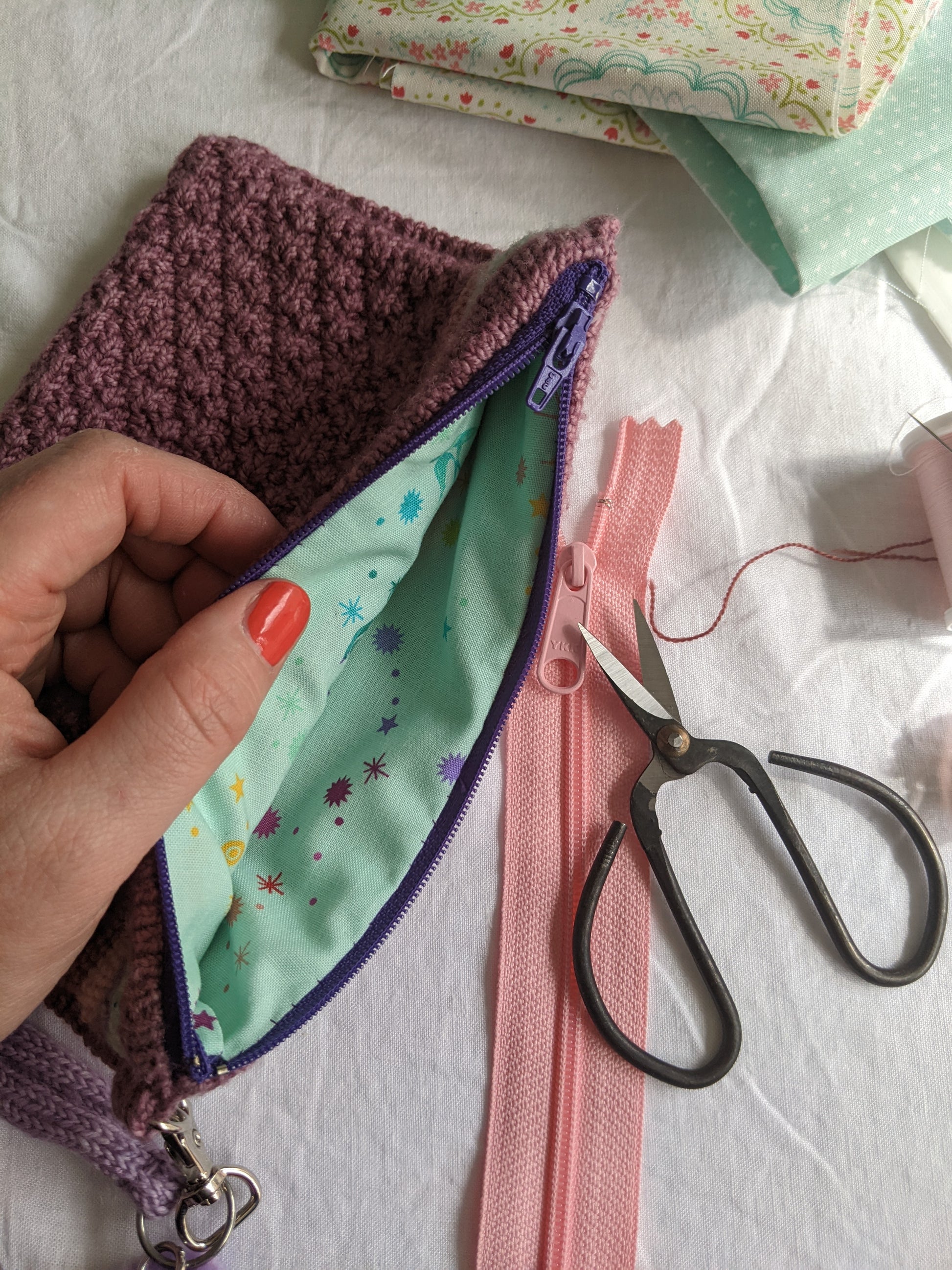 Jen's hand holds open a knit notions pouch, which lies on a table next to sewing scissors, extra zippers, and fabric. The inside of the pouch is lined with blue fabric.