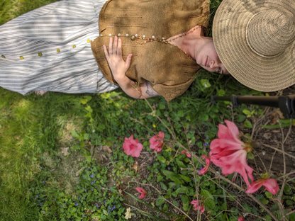 Jen lays in the grass, peeking up at the camera from under the brim of a straw hat. She wears a striped skirt with a gold dolman tee. The tee has eyelet lace strips coming up from the hem and down from the shoulder seams, and is buttoned with small pearl buttons.