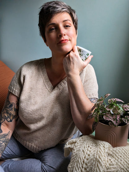 Jen wears her Classic LBD - a light beige, knit tee - with blue jeans. She leans against a table covered in a knit blanket and smiles at the camera.