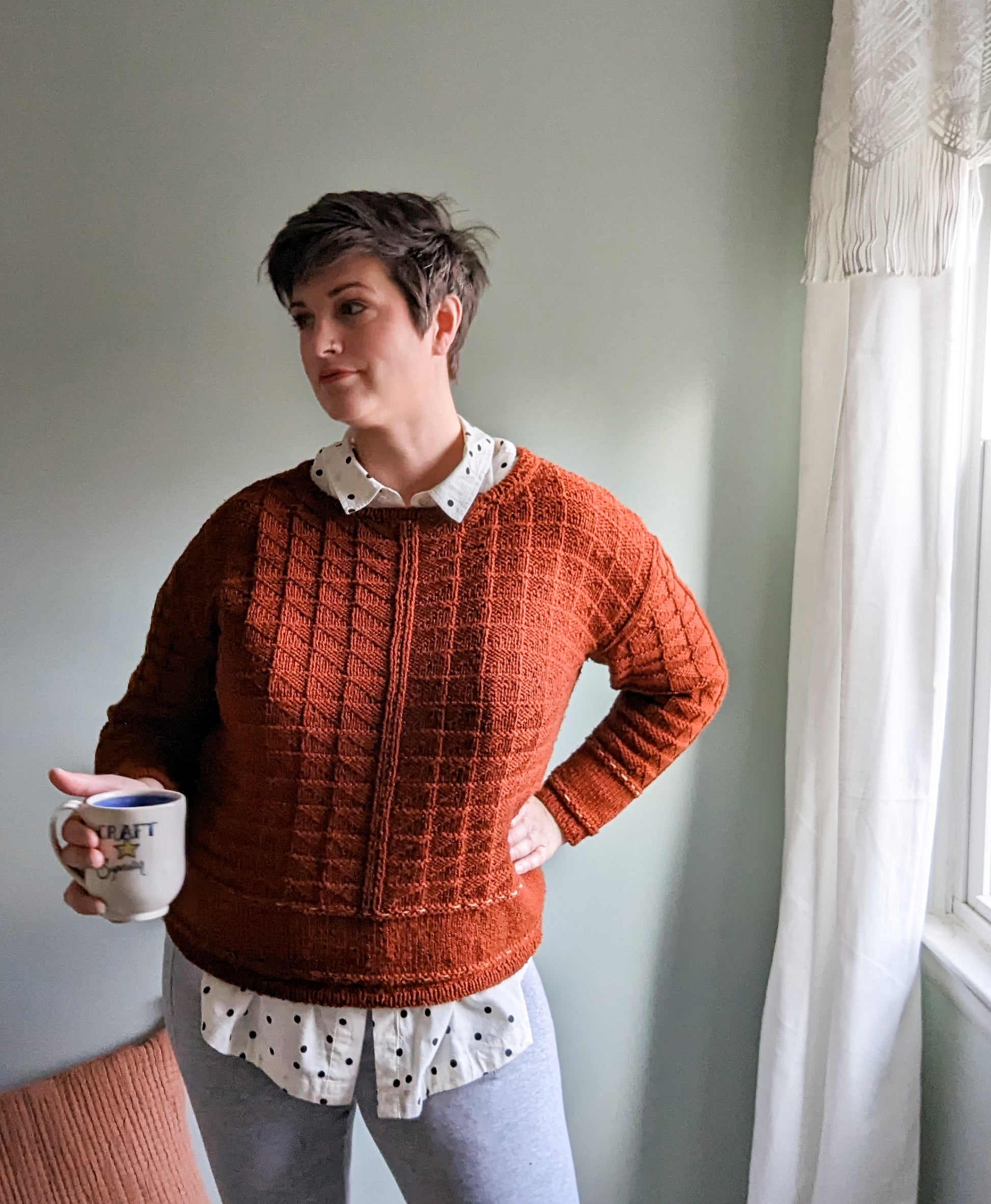 Jen holds a mug, looking off camera. She wears a brick red pullover, knit in a quilt stitch, over a polka dot button down.
