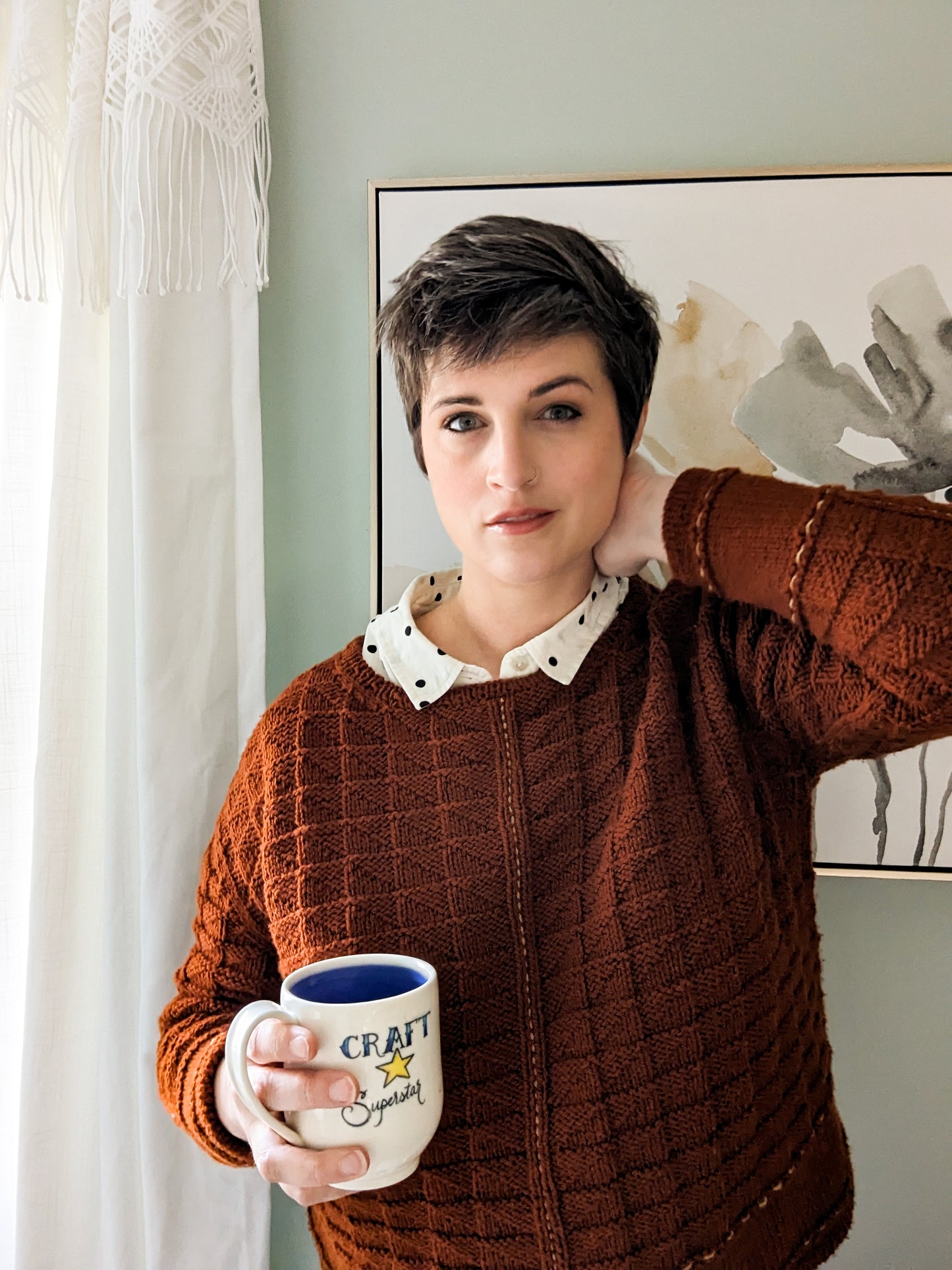 Jen holds a mug, looking at the camera. She wears a brick orange sweater, knit with an allover texture, over a polka dot button up. 