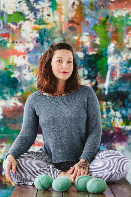 Elizabeth sits on a table cross legged. She is wearing a knit seamless tee. The knitting pattern for this tee is top-down and seamless.