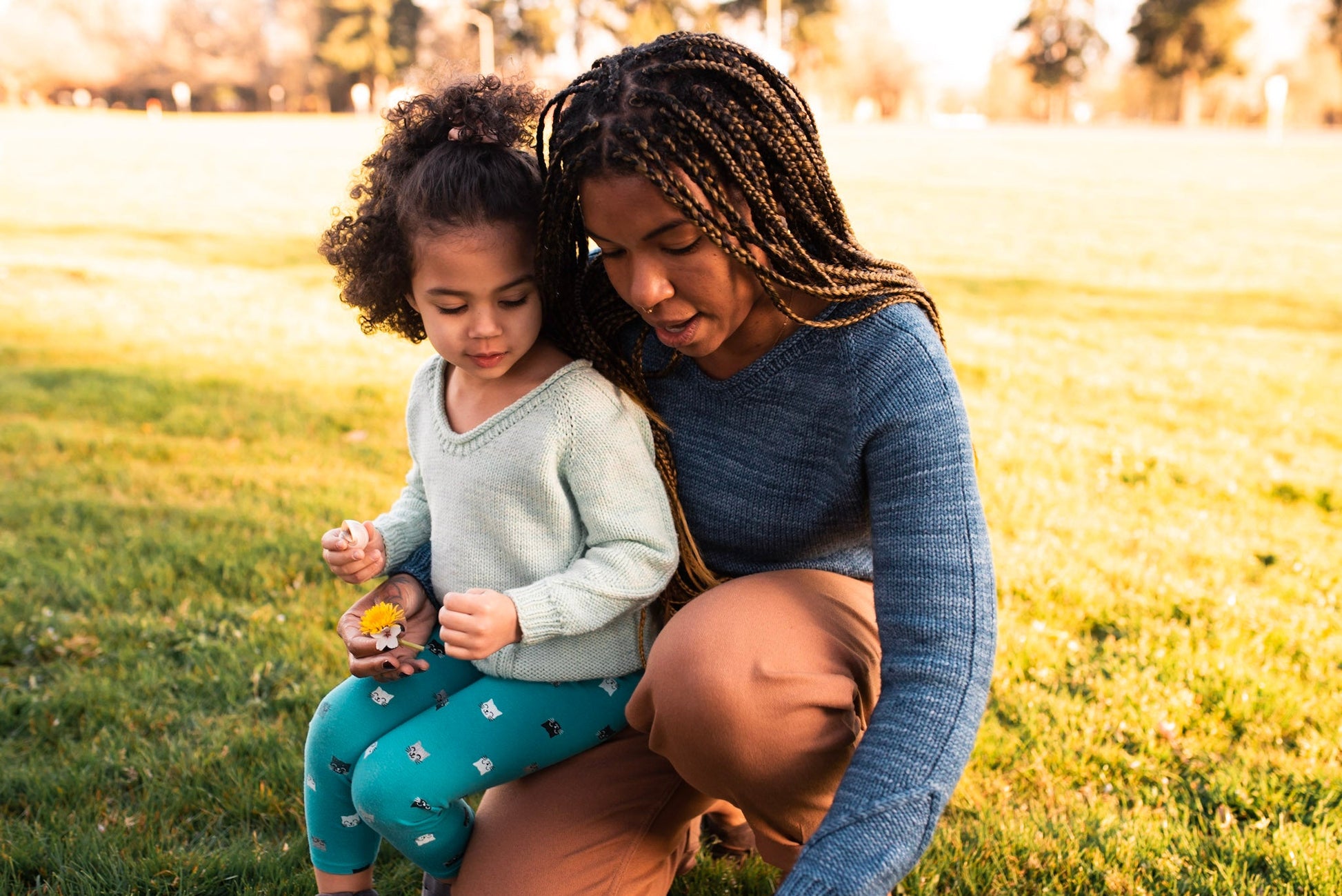 Rachel holds Mia on her knee as she shows her something in the grass. They wear matching hand knit sweaters, each with a scoop neck and cabling details around the cuffs.