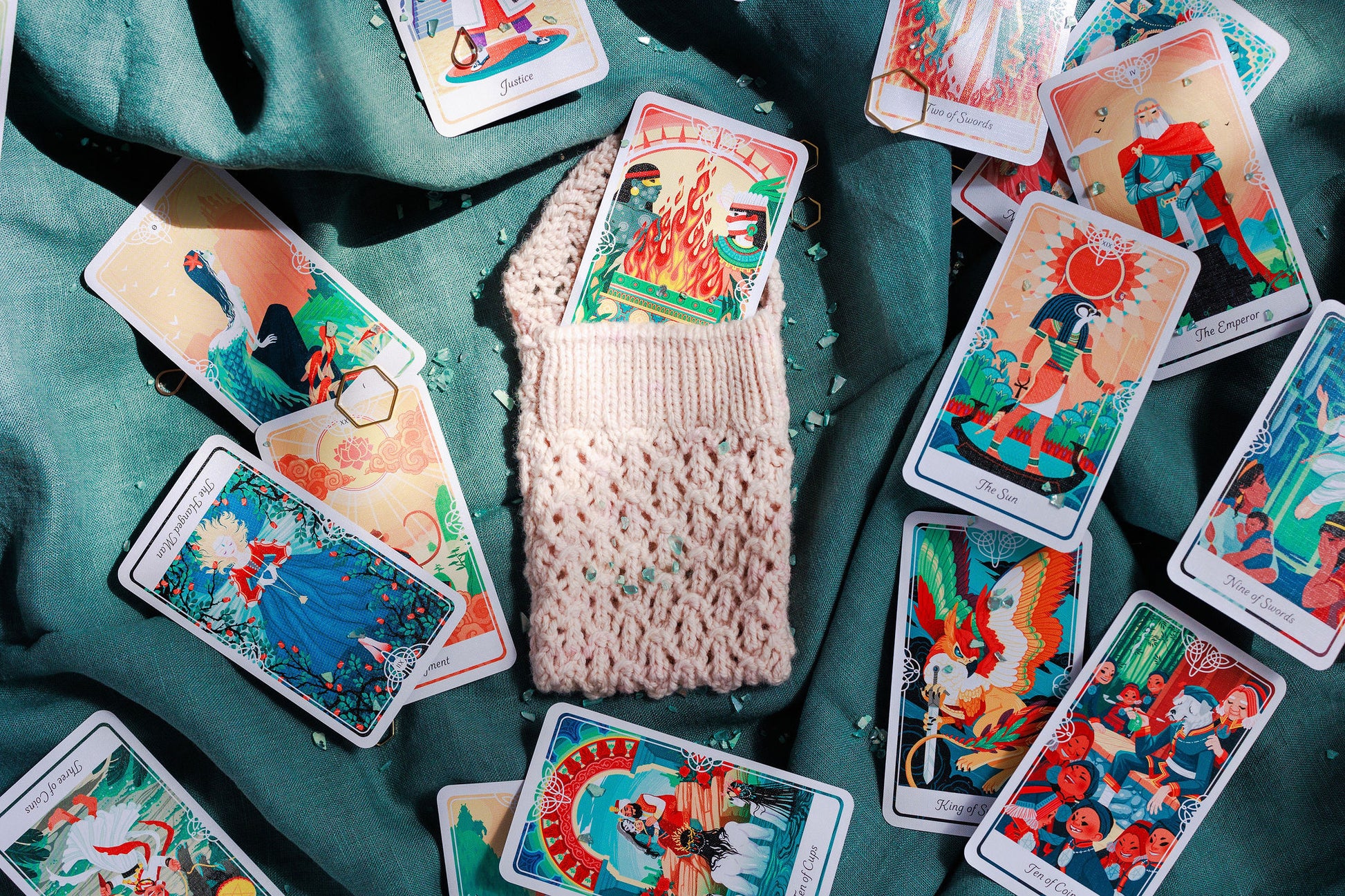 A cream lace knit pouch (the Mary Tarot ouch) lays on a green fabric background. Inside and around the pouch are scattered colorful tarot cards.