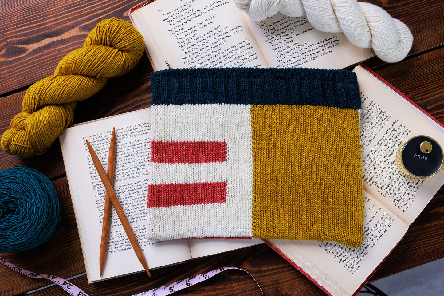 A pouch, the Case Study pouch, lies on a table surrounded by books, yarn, and knitting needles. The pouch features an intarsia colourblock design, made up of yellow, white, red, and blue sections.