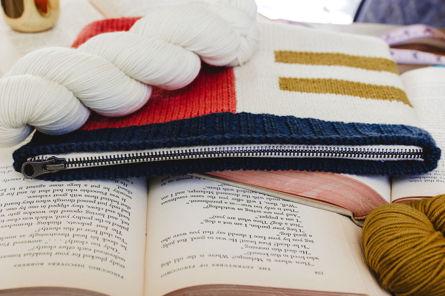 A hand knit notions case, zipped close, lies on top of open books, under a cream skein of yarn. The case is knit in a colorblock design, using intarsia techniques, made with cream , ochre, pink, and navy blue yarn.