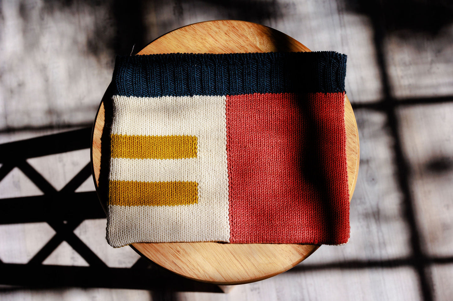 A notions case, knit with navy blue, pink, ochre, and cream yarn in an intarsia design, sits on a wooden stool.