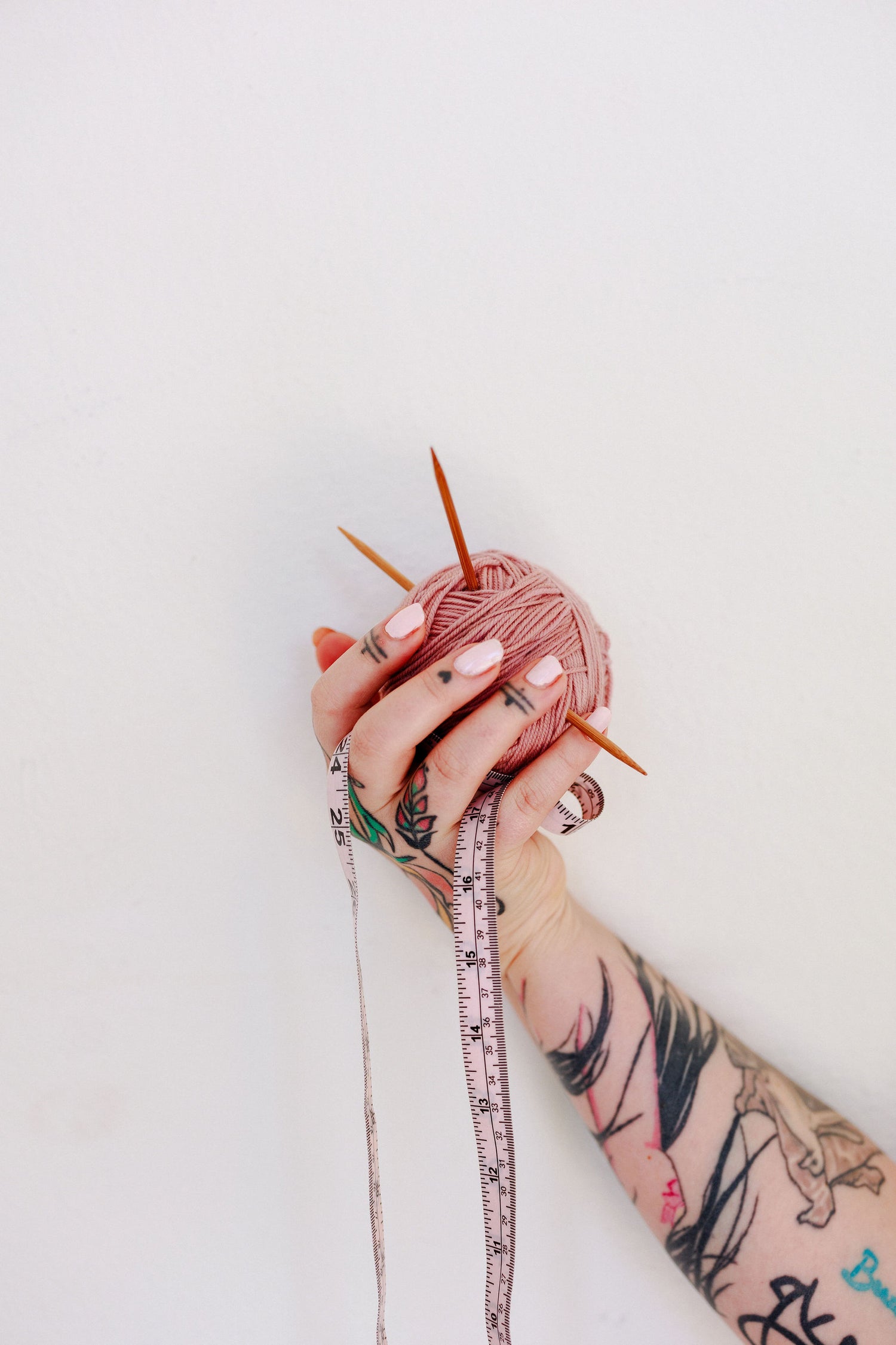 A woman's arm with tattoos holds a ball of pink yarn with knitting needles. A dressmaker's tape measure is spooled in her hand and drapes towards the bottom of the frame.