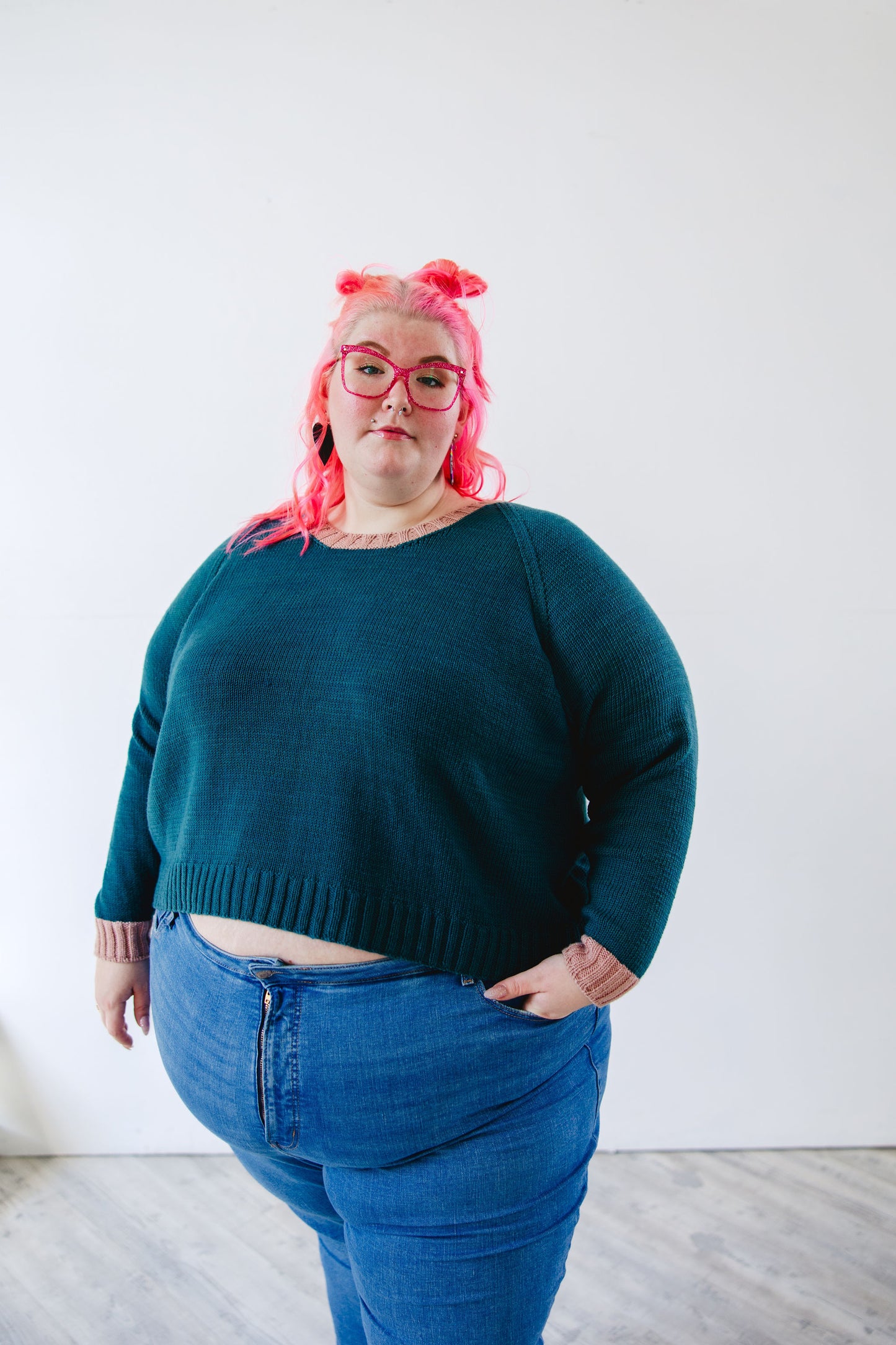 Courtney stands, looking at the camera, wearing a teal sweater knit with pink accents at the cuffs and neckband. Her pink glasses match her pink hair.