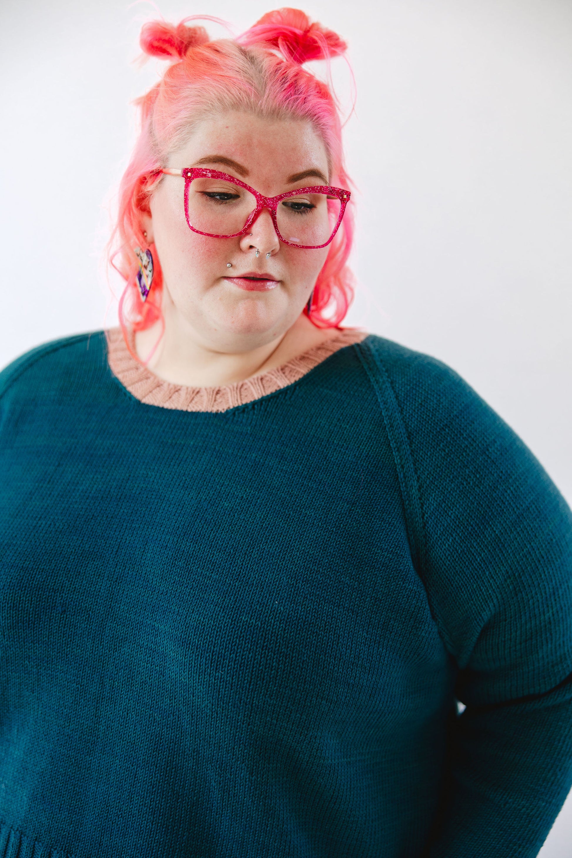 Seen close up, Courtney wears a teal hand knit sweater with an pink neckband. She looks off camera, her pink glasses matching her pink hair.