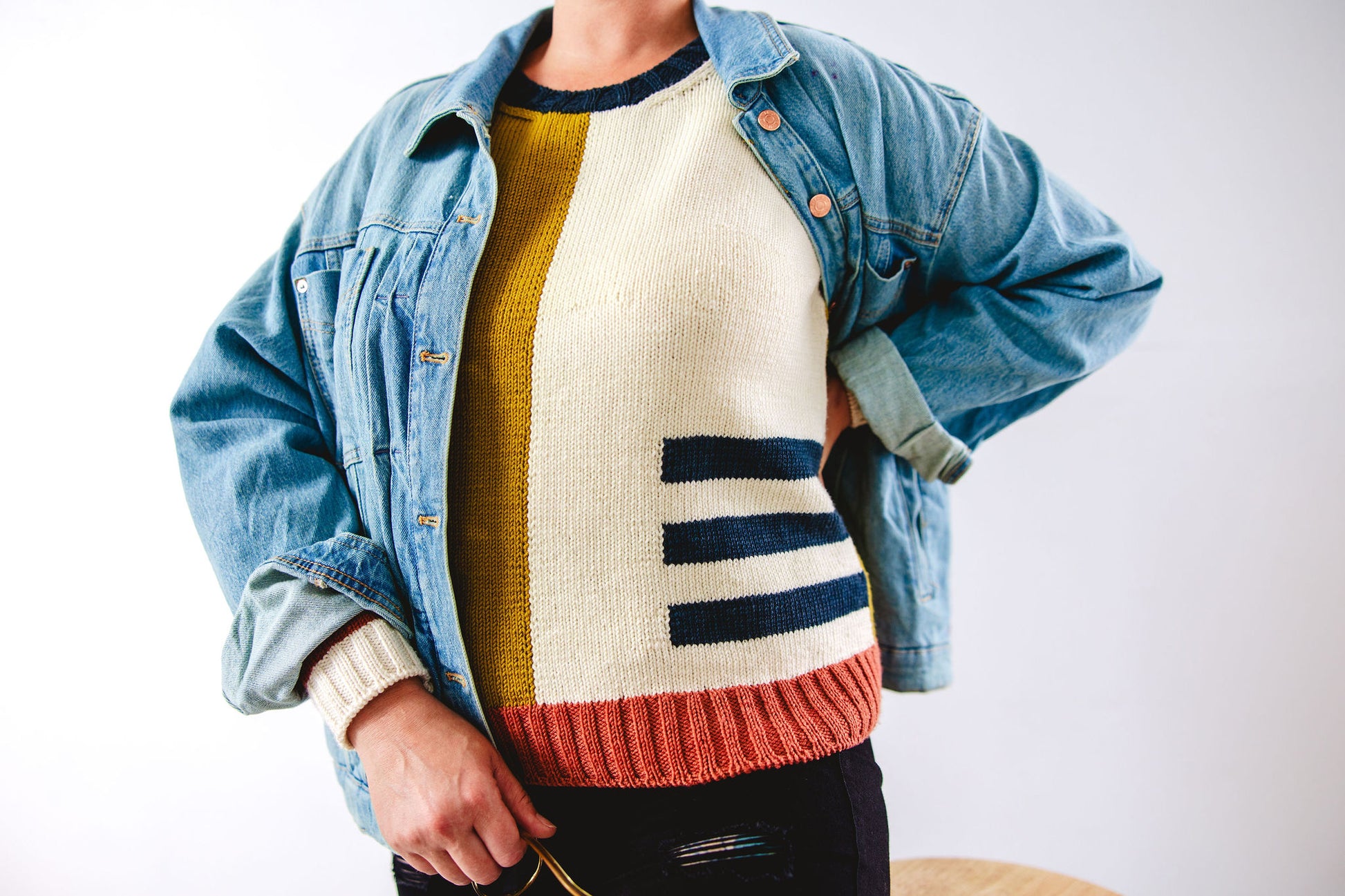 Seen from the shoulders down, Jen wears a denim jacket on top of her hand knit sweater. The sweater uses intarsia techniques to create color blocks of ochre, white, navy blue, and pink.