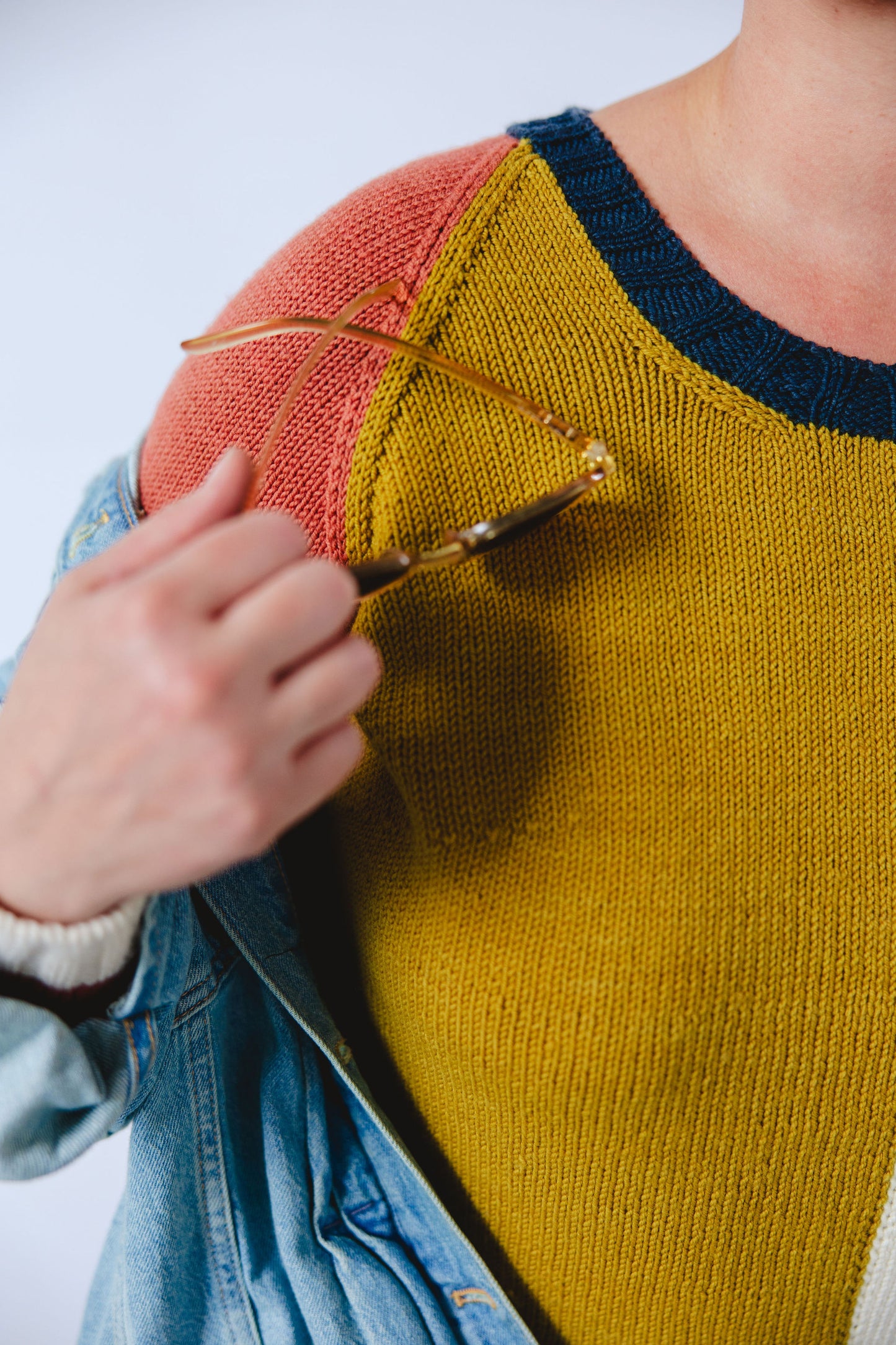 Seen close up, Jen holds a pair of sunglasses near her shoulder, in the act of removing her denim jacket. The camera focus on the details of her hand knit sweater, made up of navy blue, white, ochre, and pink color blocks.