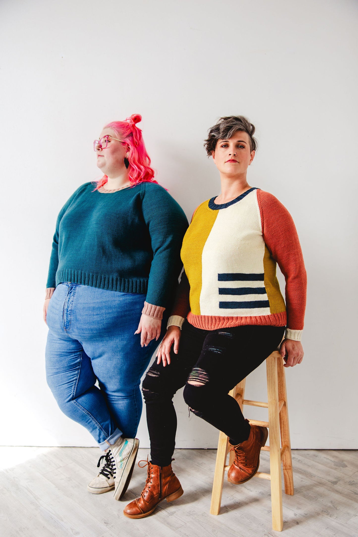 Courtney, who wears a solid green version of the Study Group pullover with contrasting pink cuffs, stands next to Jen and looks off camera. Jen is seated on a stool next to Courtney, wearing a yellow, white, red, and blue version of the Study Group pullover, knit with intarsia techniques.