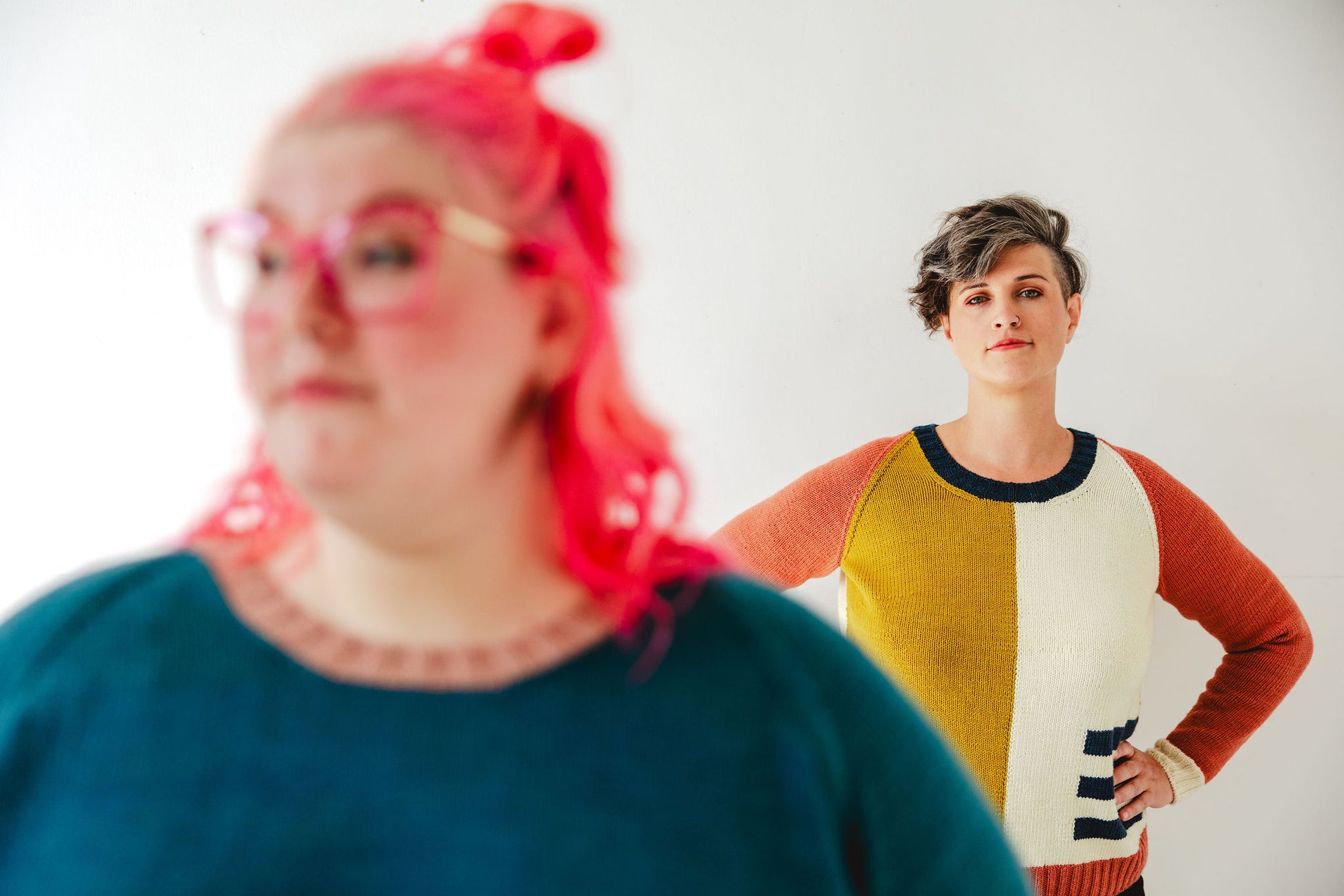 Jen stands behind Courtney, the camera focused on her. She wears an intarsia knit sweater of white, navy blue, pink, and ochre. In front of her, out of focus, Courtney wears a teal sweater knit with a pink, ribbed neckband.