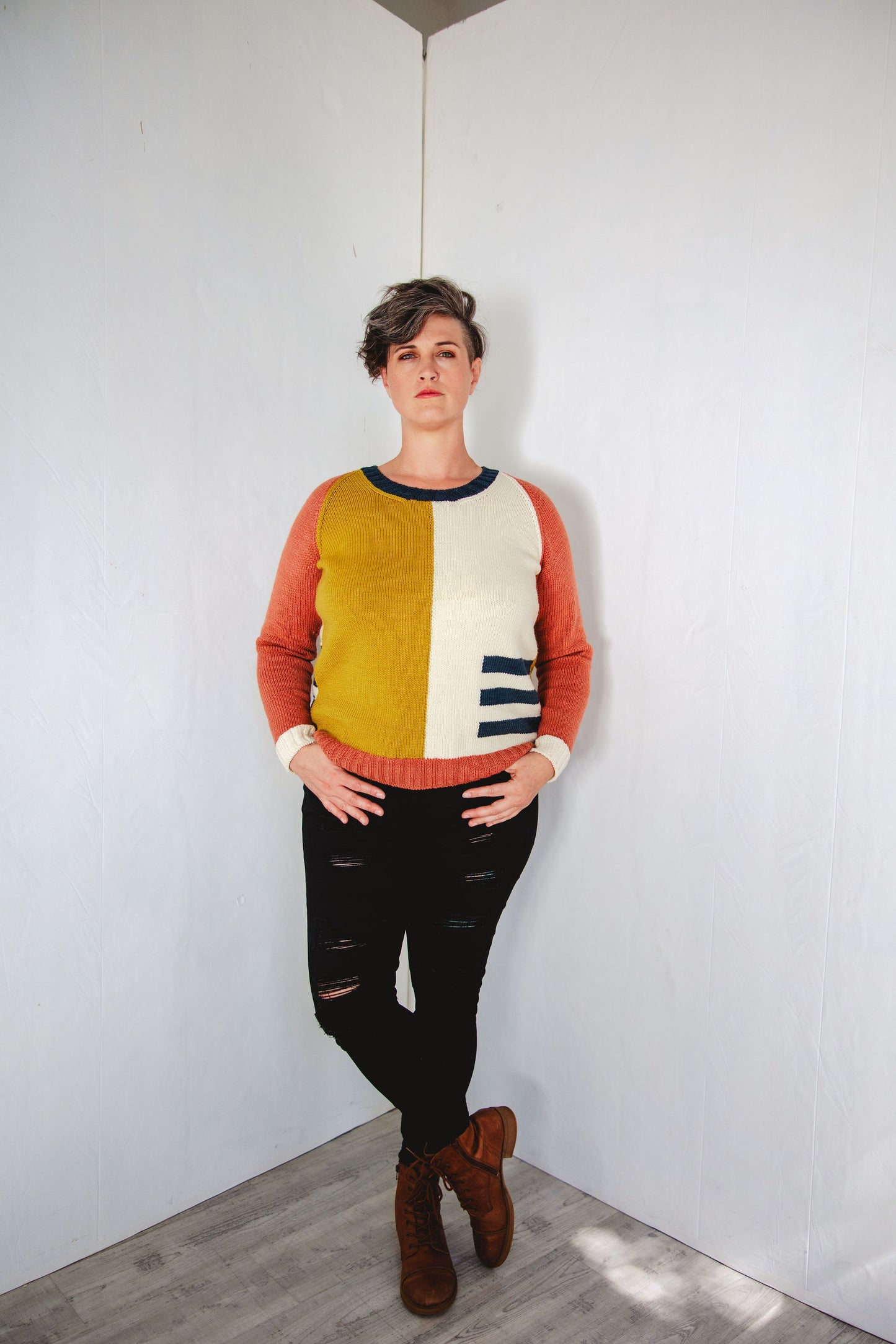 Jen stands, hands in the pockets of her black jeans, looking at the camera. Her hand knit sweater features color blocks of ochre, white, navy blue, and pink, knit using intarsia techniques.