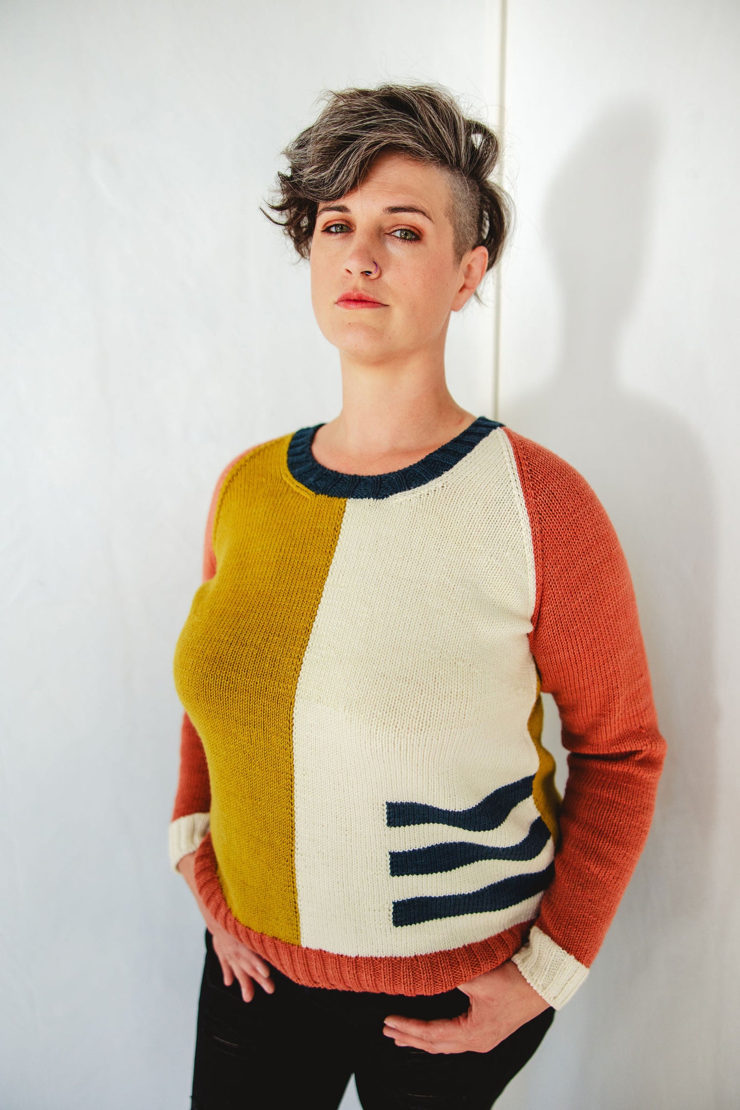 Jen stands against a white wall, looking towards the camera. She wears a yellow, white, red, and blue colorblock knit sweater (the Study Group sweater) with black pants.