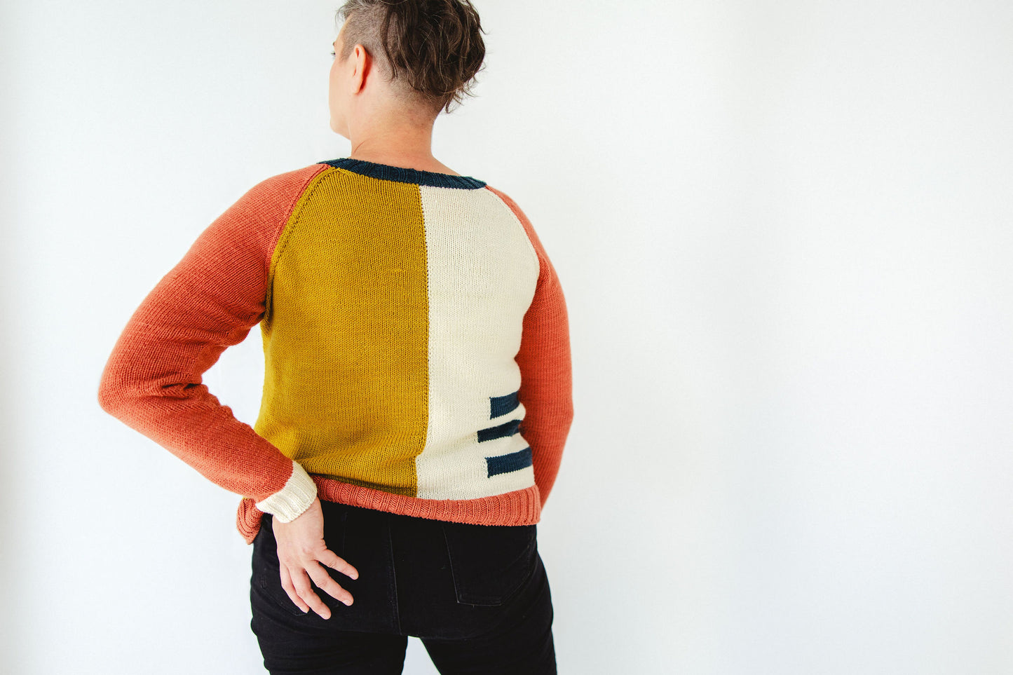 Jen is seen from behind, wearing a yellow, white, blue, and red knit sweater with an intarsia colorblock design. She also wears a pair of black jeans.
