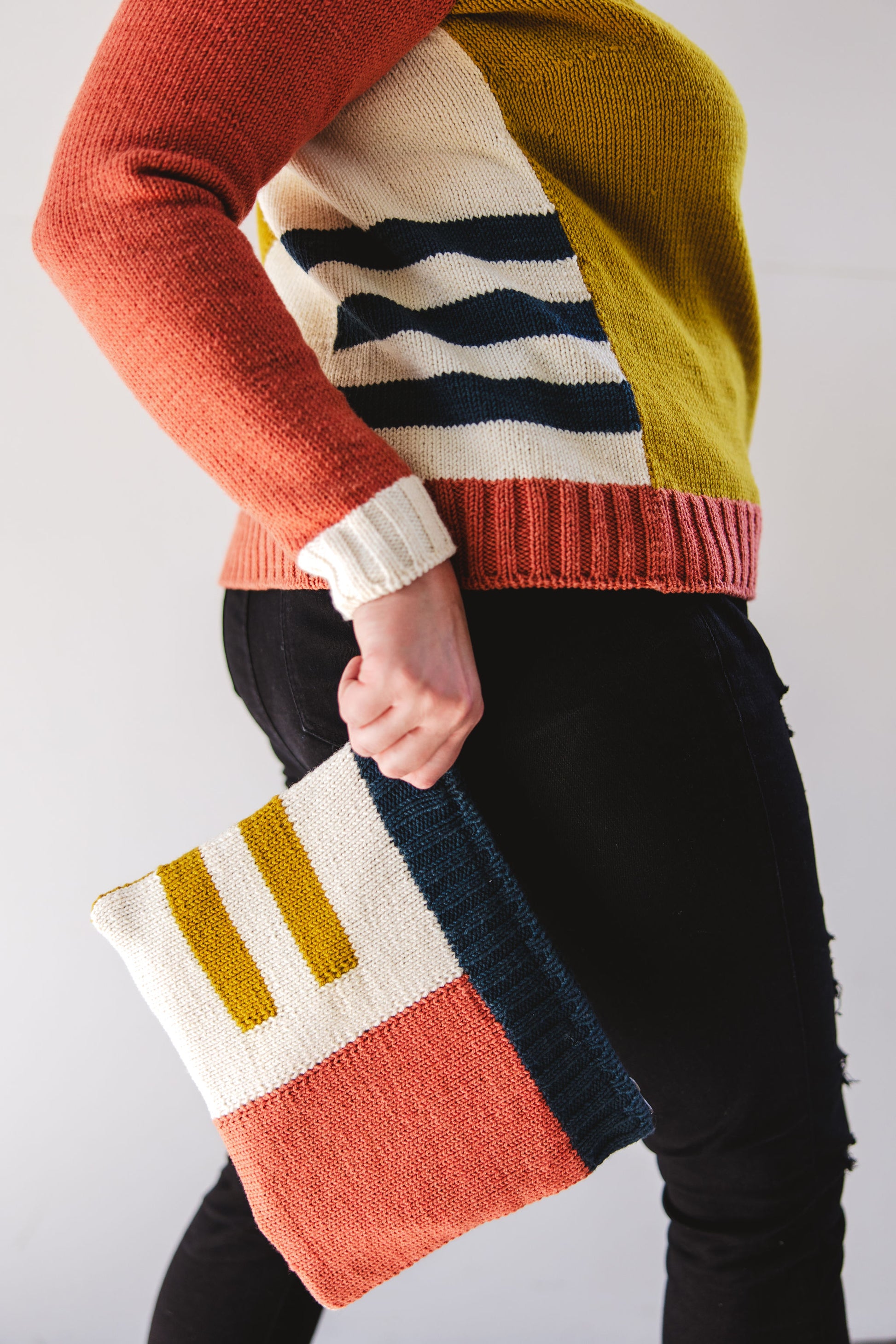 Seen from the shoulders down from the side, Jen holds a hand knit notions case, made from cream, ochre, pink, and navy blue yarn in a colorblock intarsia design. Jen wears a matching sweater, in the same colors and pattern, with black jeans.