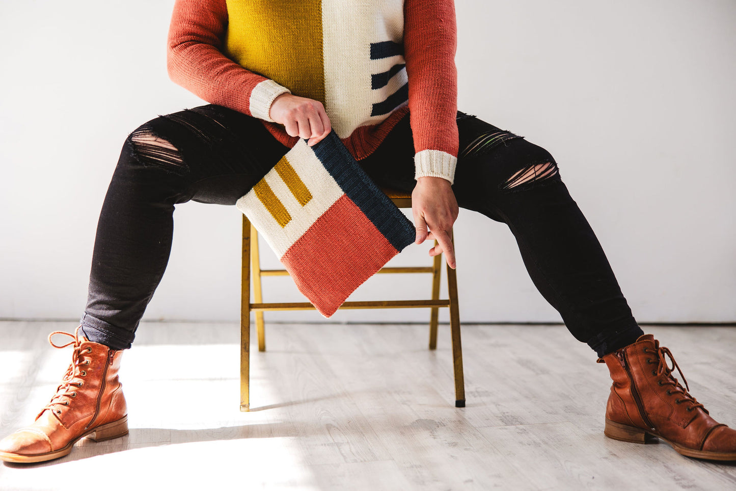 Seen from the shoulders down, Jen sits on a chair, holding a notions case, knit in pink, ochre, navy blue, and cream intarsia. She wears a sweater, knit in the same intarsia design in matching colours, with black ripped jeans and brown boots.