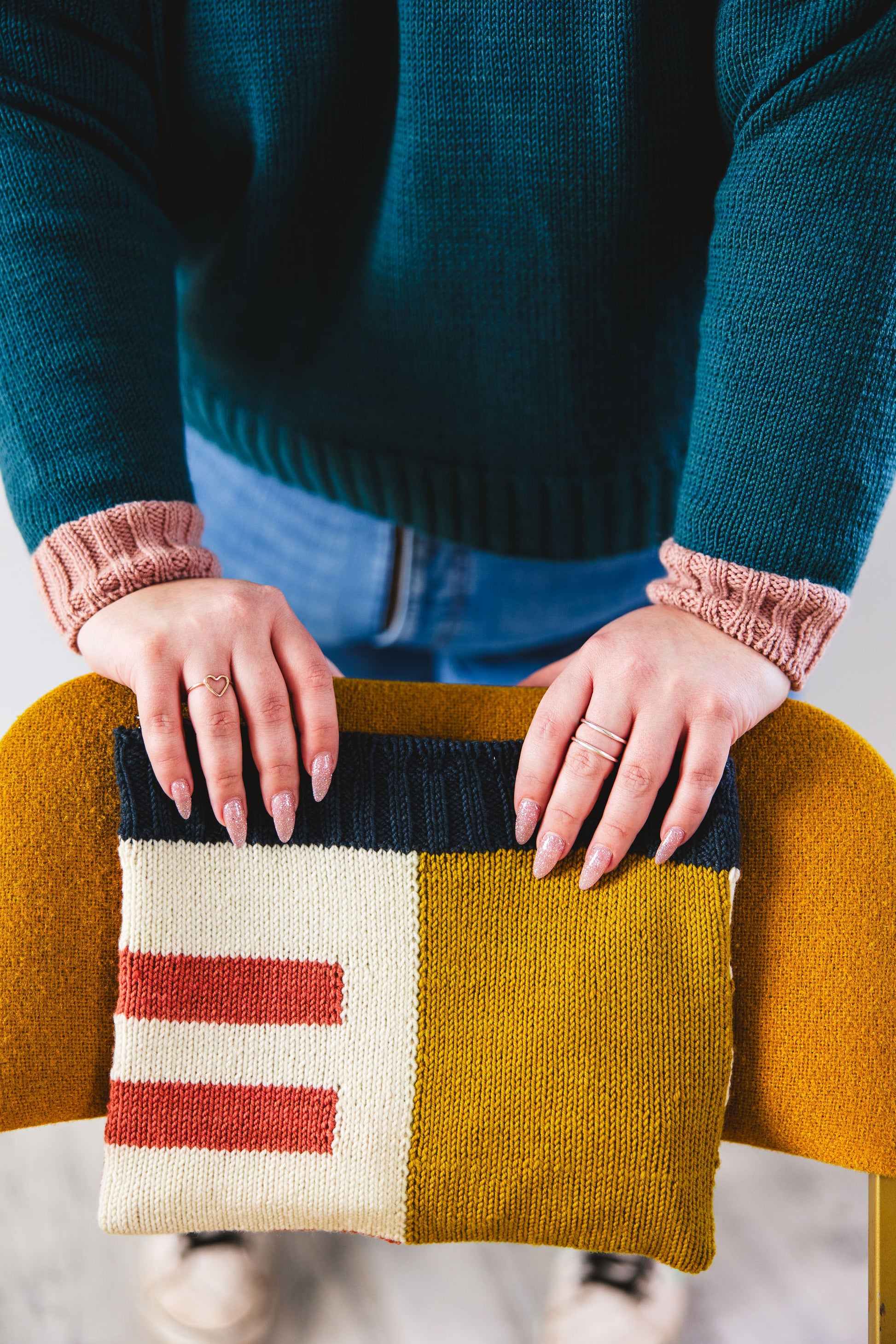 Courtney, who wears a green pullover with pink cuffs knit from the Study Group pattern, is seen from the torso down. She holds a knit pouch, the Case Study pouch. The pouch has a white, red, yellow, and blue colourblock design, created using intarsia techniques.