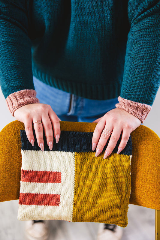 Seen from the shoulders down, Courtney holds an intarsia knit notions case against the back of a chair. The case is knit with ochre, cream, pink, and navy blue yarn. Courtney wears a teal sweater, knit with contrasting pink cuffs.