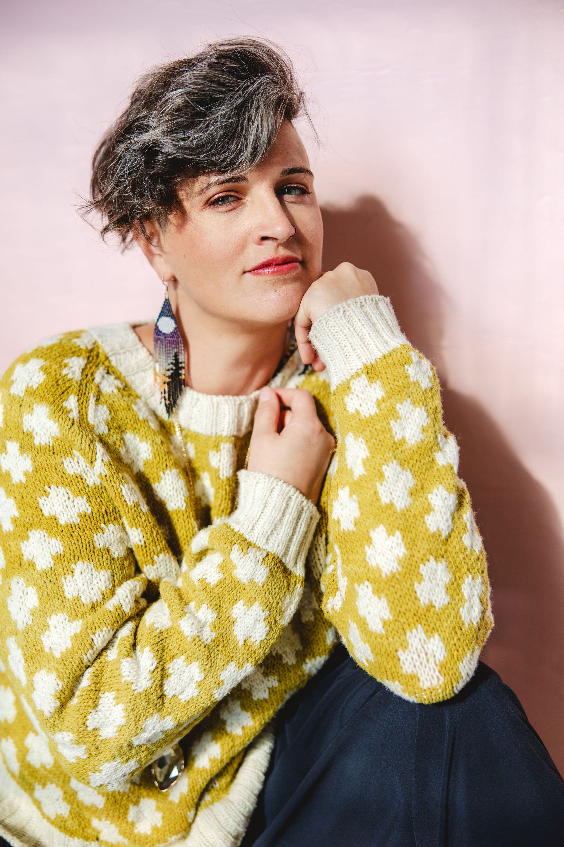 Jen kneels and smiles at the camera. She wears a yellow knit sweater with a white plusses pattern and trim.