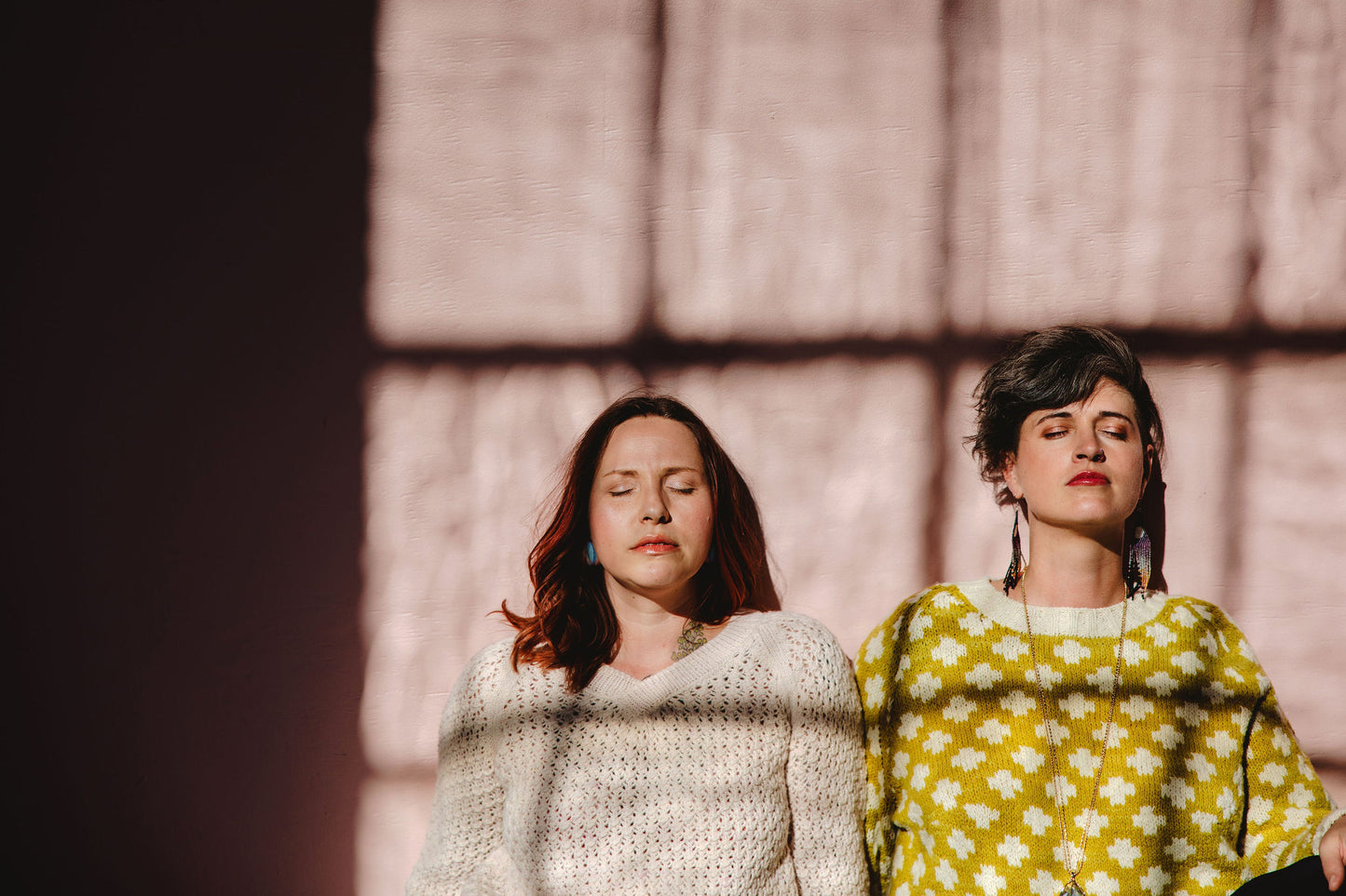 Bess and Jen stand against a wall, seen from the waist up, with their eyes closed. Bess wears a cream lace knit sweater (the Mary Raglan), and Jen wears a yellow knit sweater with a white plusses pattern and trim (the Plusses sweater).