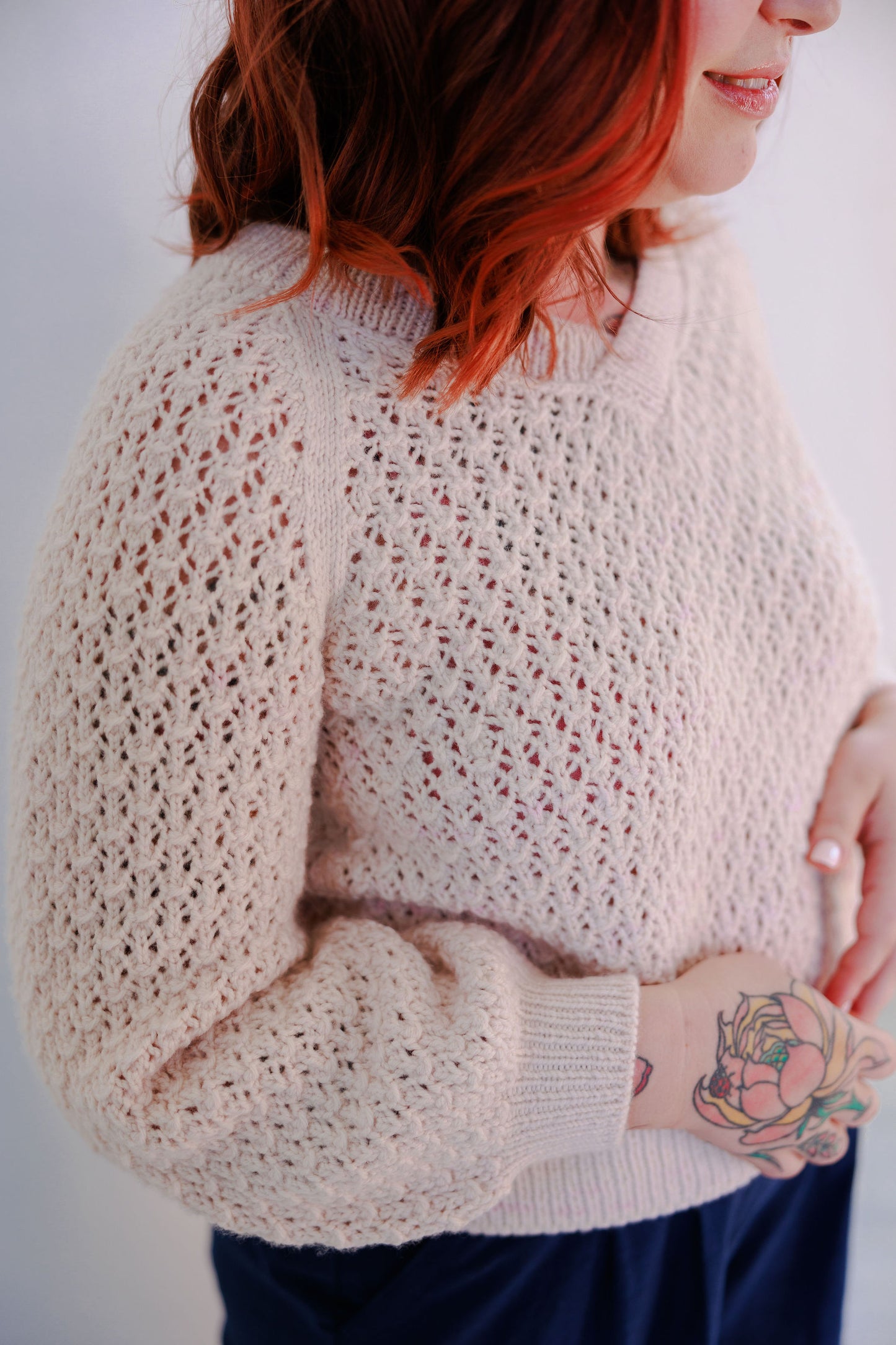 Bess wears a cream lace knit sweater, the Mary Raglan. She is seen at a 3/4 angle, and is smiling.