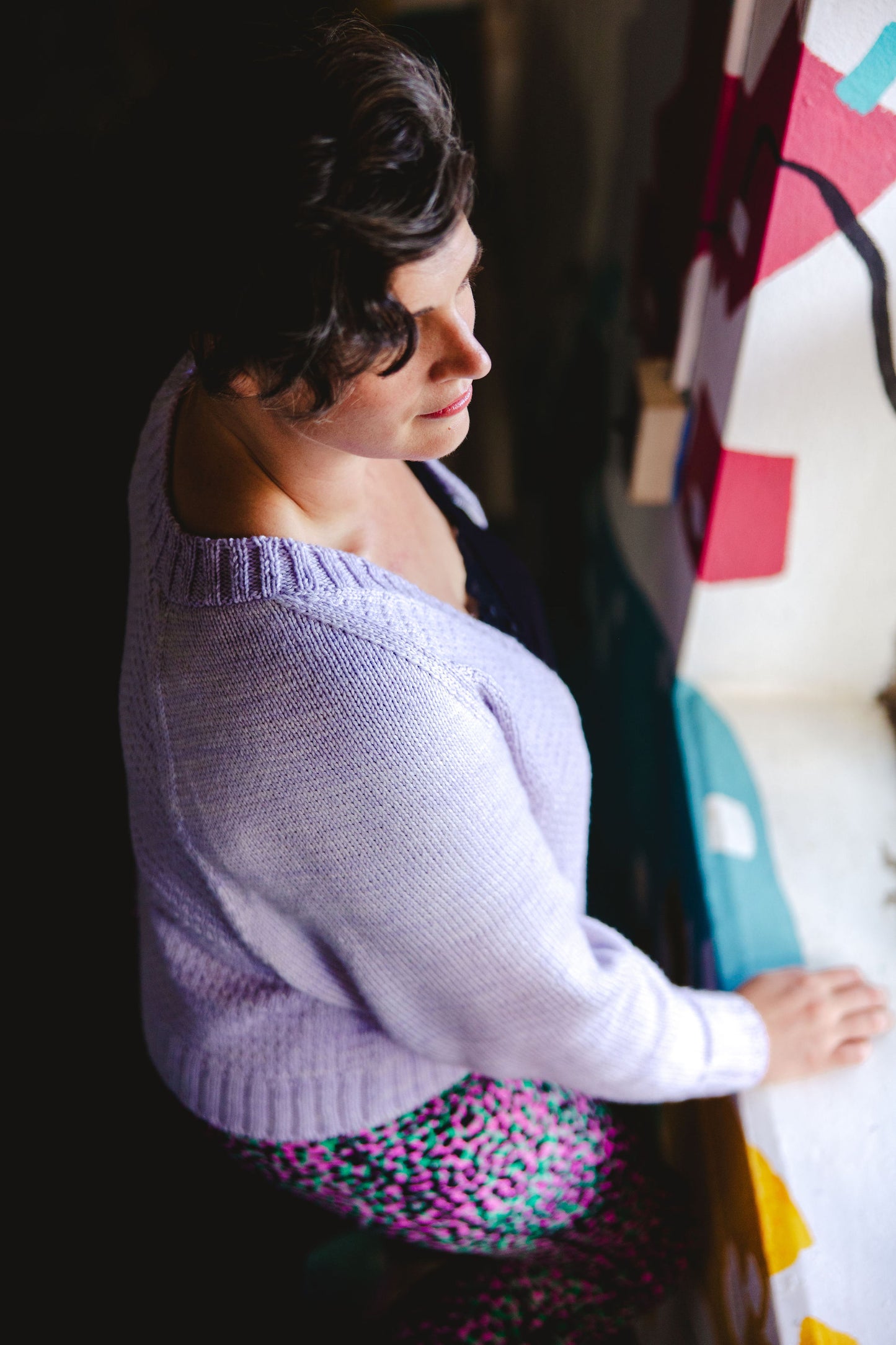 Seen from above, Jen stands in front of a window, wearing a light purple knit sweater, partially zipped over a black camisole.