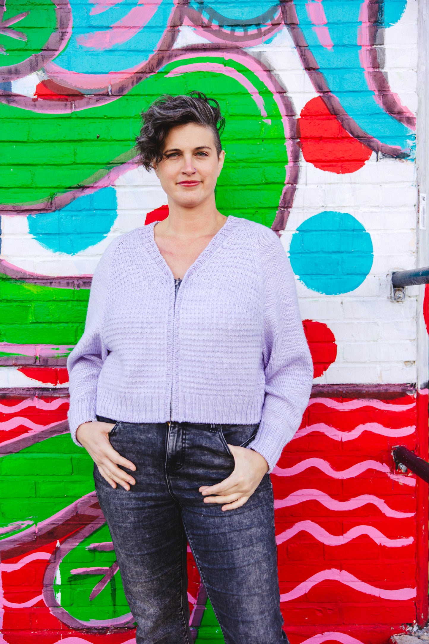 Jen stands in front of a wall mural, smiling at the camera and wear a light purple zip sweater with acid wash black denim jeans. The sweater is knit up in a simple seed stitch and a ribbed V-neckline.