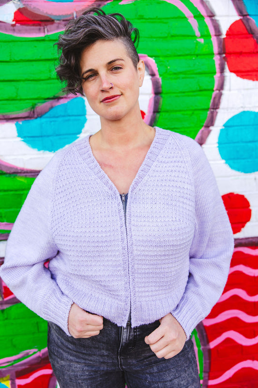 Jen stands against a wall mural, smiling at the camera. She wears dark acid wash jeans with a light purple zip-up sweater, knit with a V-neck line and a subtle seed stitch.