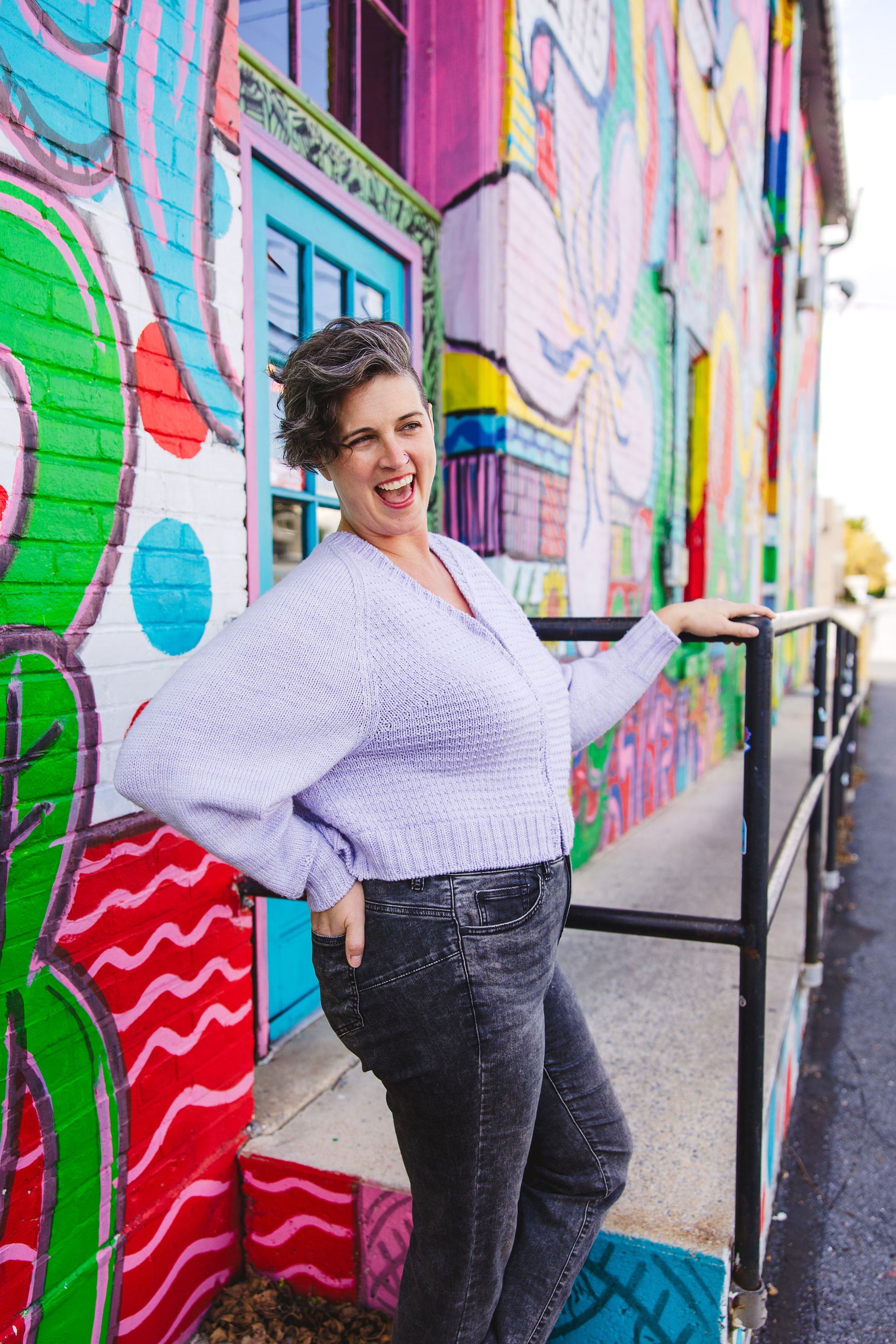 Jen, wearing a light purple zip-up sweater, holds the railing of a ramp in front of a wall mural and smiles, looking off camera. She has one hand on her hip, showing off the roomy sleeves on the sweater, which she wears with black acid wash jeans. A wall mural can be seen in the background.