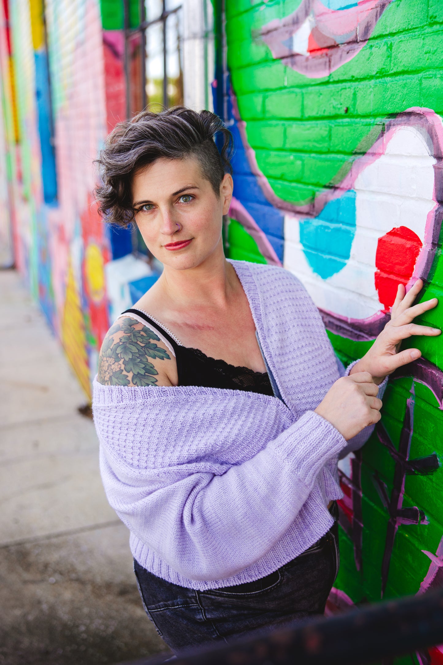 Jen, standing next to a wall mural, looks past the camera. She wears a light purple knit sweater, partially unzipped so it falls off one shoulder revealing a black camisole.