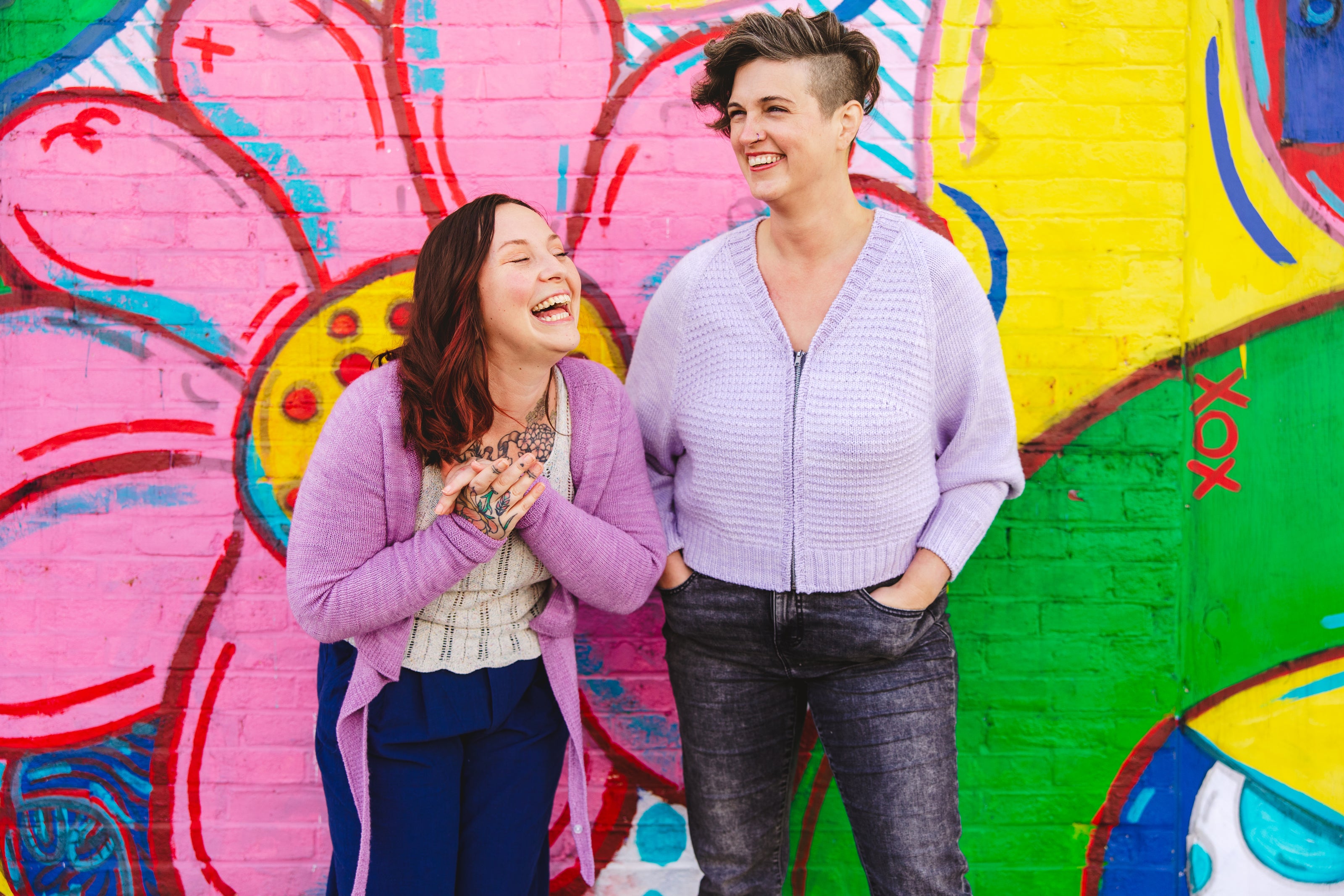 Jen and Bess stand in front of a wall mural, laughing. Bess wears a lace knit cream tank top under a purple-pink knit ballet wrap with a pair of navy pants. Beside her, Jen wears a zip knit sweater, knit with purple yarn in a seed stitch pattern and a V-neckline, along with acid wash black denim jeans.